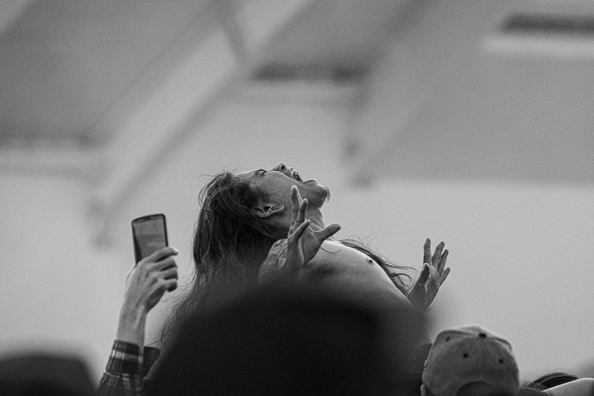Black and white photo of a woman singing with her head tilted back, eyes closed, and one hand raised, in a crowd with someone holding a phone