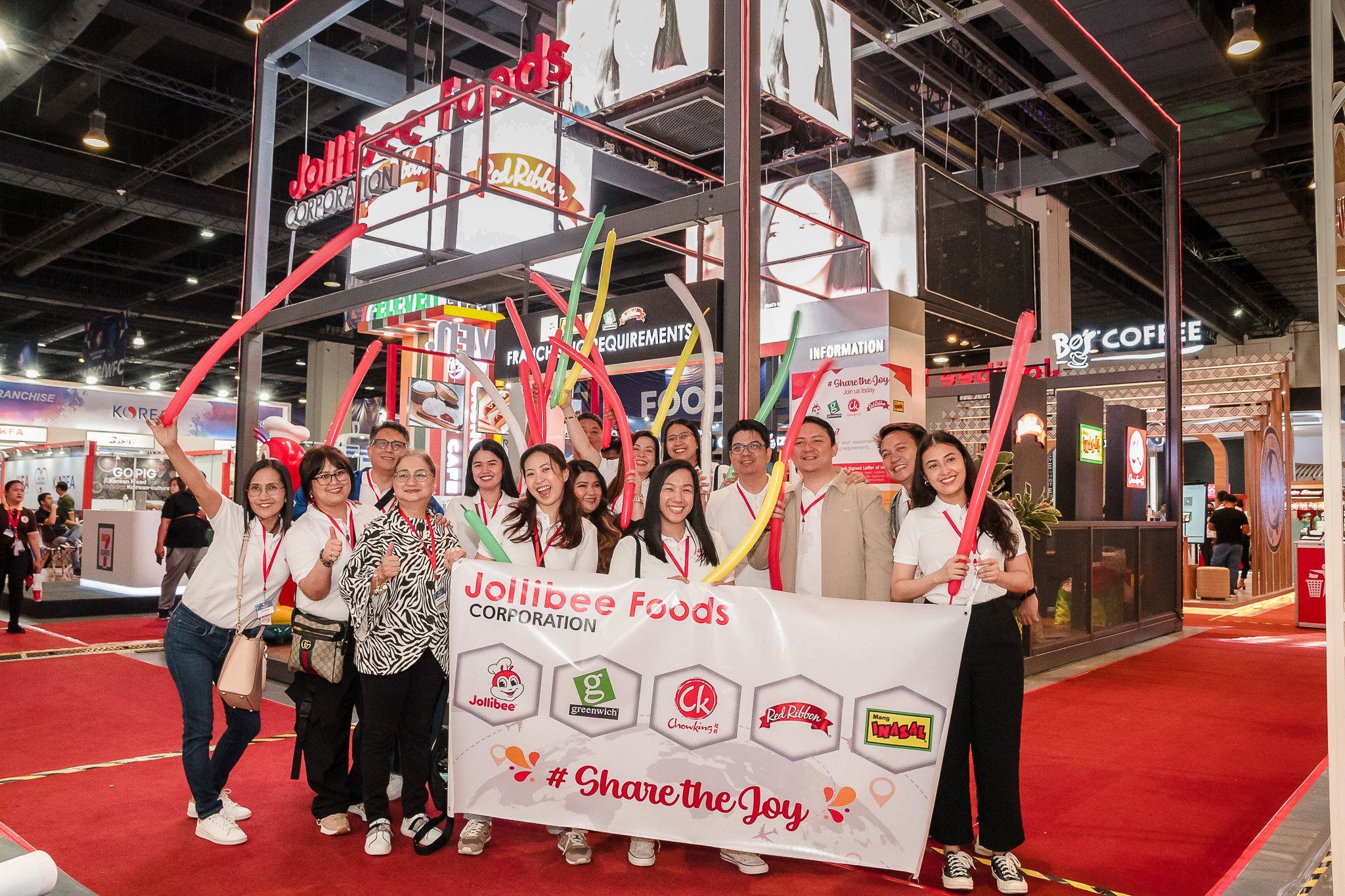 Group of people holding balloons and a Jollibee Foods Corporation banner at an indoor event, with various food booths and signage visible in the background.