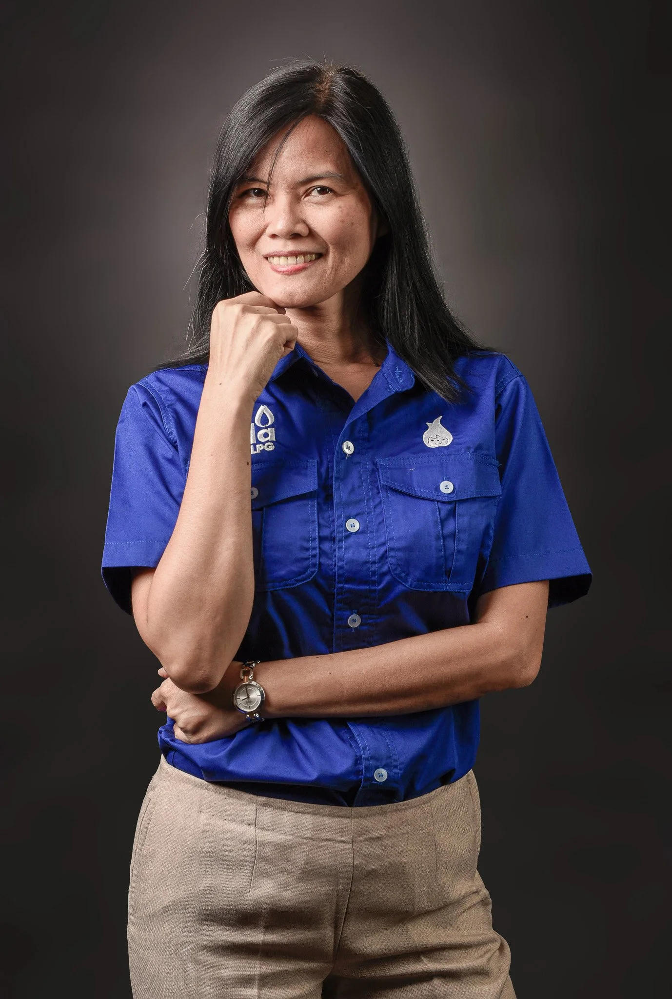 A woman with black hair wearing a blue short-sleeved button-up shirt and beige pants, standing against a dark background with a confident smile.
