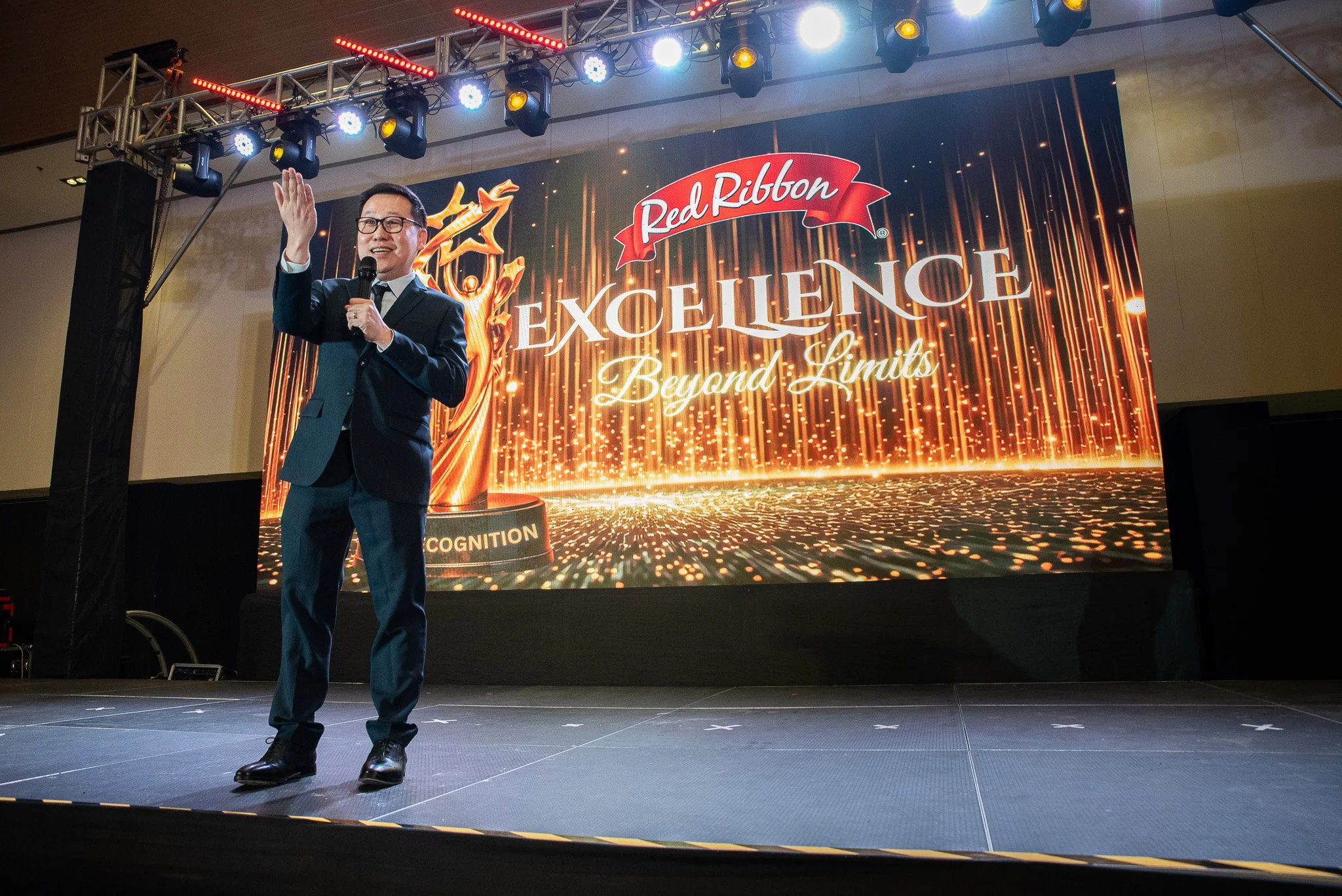 A man in a suit holding a microphone speaking on a stage with a large display behind him that says 'Red Ribbon Excellence: Beyond Limits' and features a golden trophy with red ribbon details.