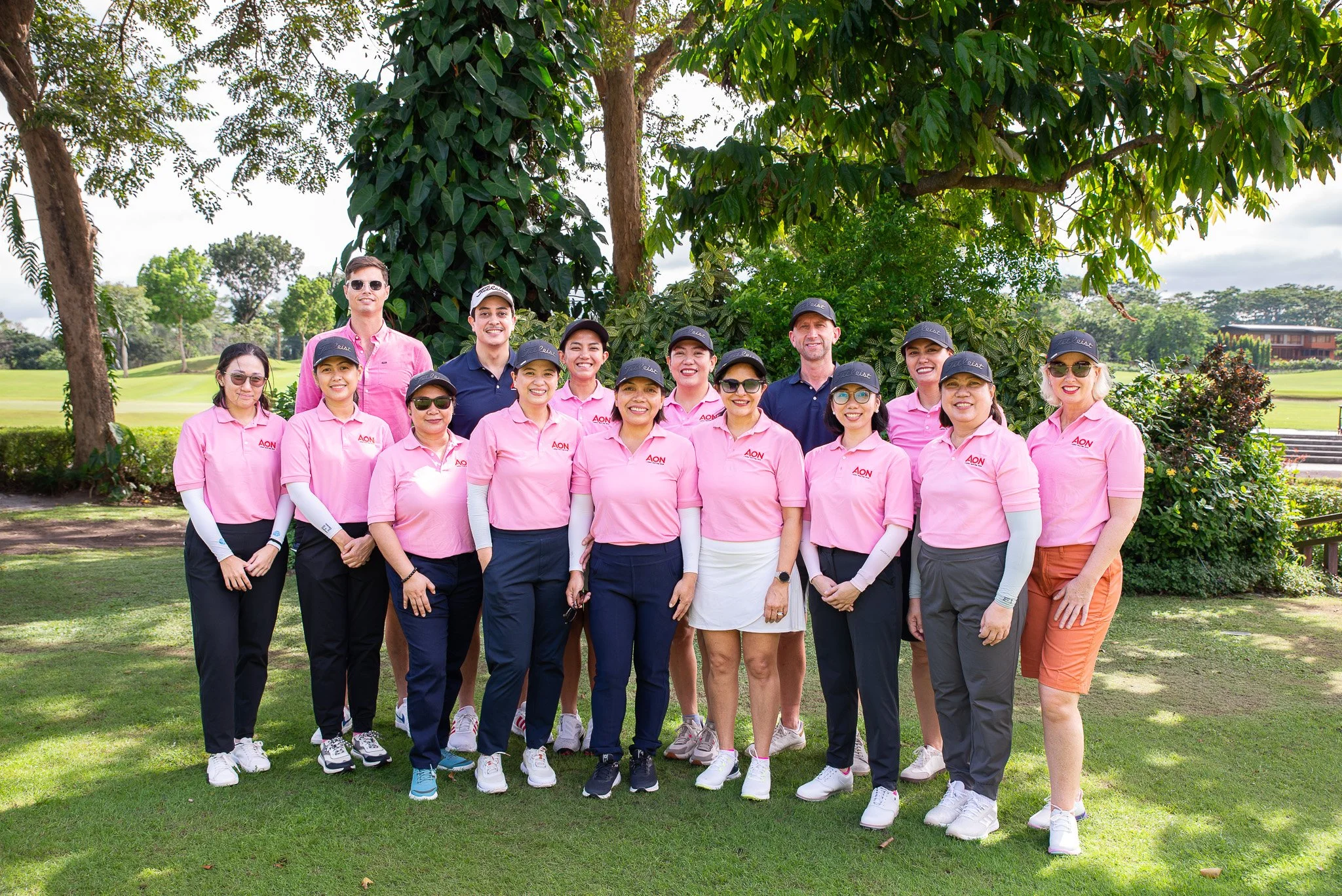 Group of people posing on a golf course, wearing pink polo shirts with 'AON' logo, some with caps and sunglasses, standing in front of a large tree and grassy field under a partly cloudy sky.