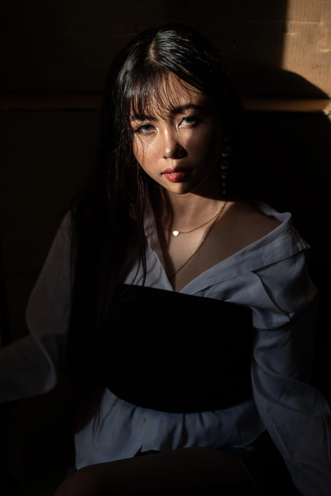A young woman with long dark hair and light skin, sitting with sunlight casting shadows on her face, wearing a white shirt, a gold necklace with a small heart pendant, and an earring, against a dark background.
