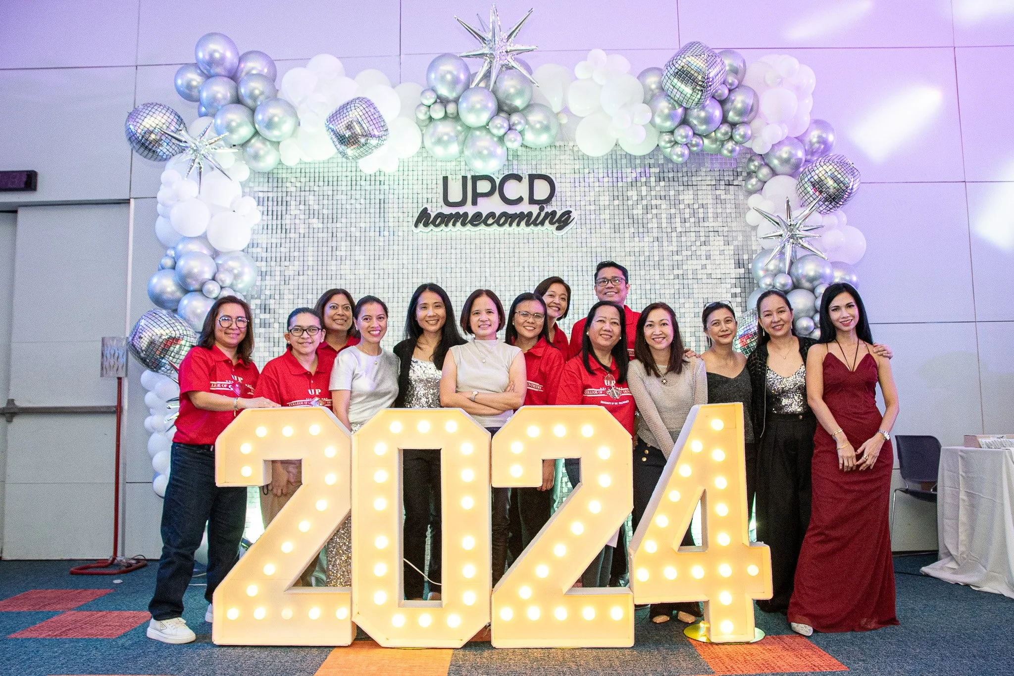 Group of people celebrating UPCD homecoming in 2024, standing behind large illuminated "2024" letters, with a festive balloon arch and mosaic backdrop.