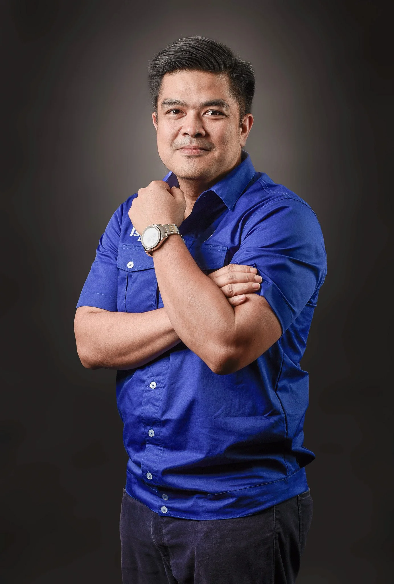 man wearing a blue short-sleeved shirt and black pants, standing with arms crossed, posing against a dark background.