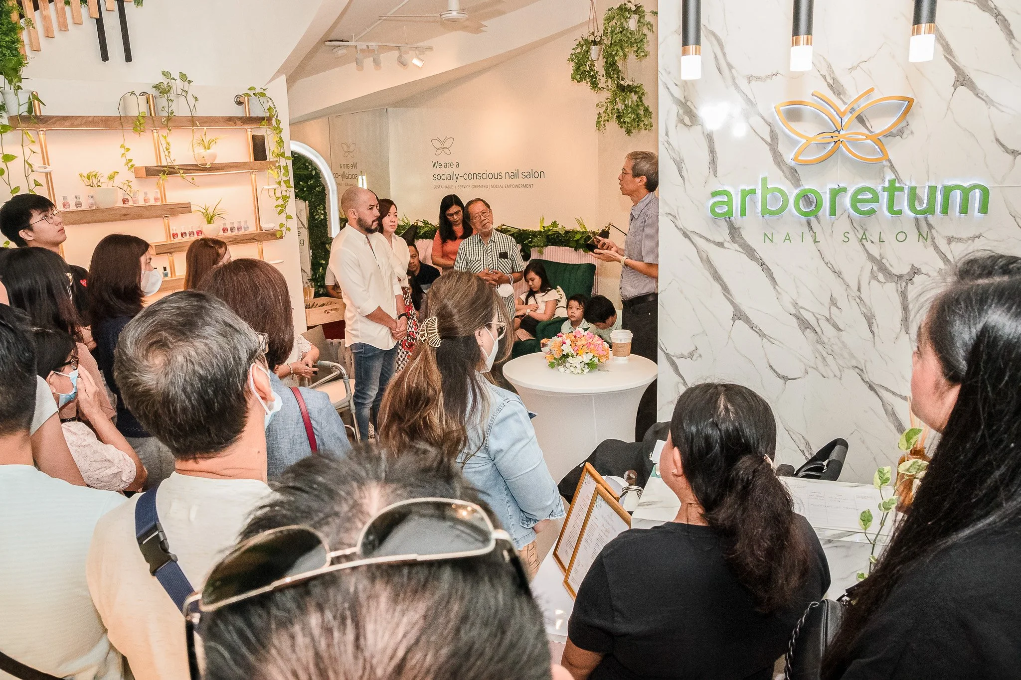 Group of people attending a presentation at Arboretum Nail Salon, with a speaker showing on a device, in a modern salon interior with plants and decorative shelves.