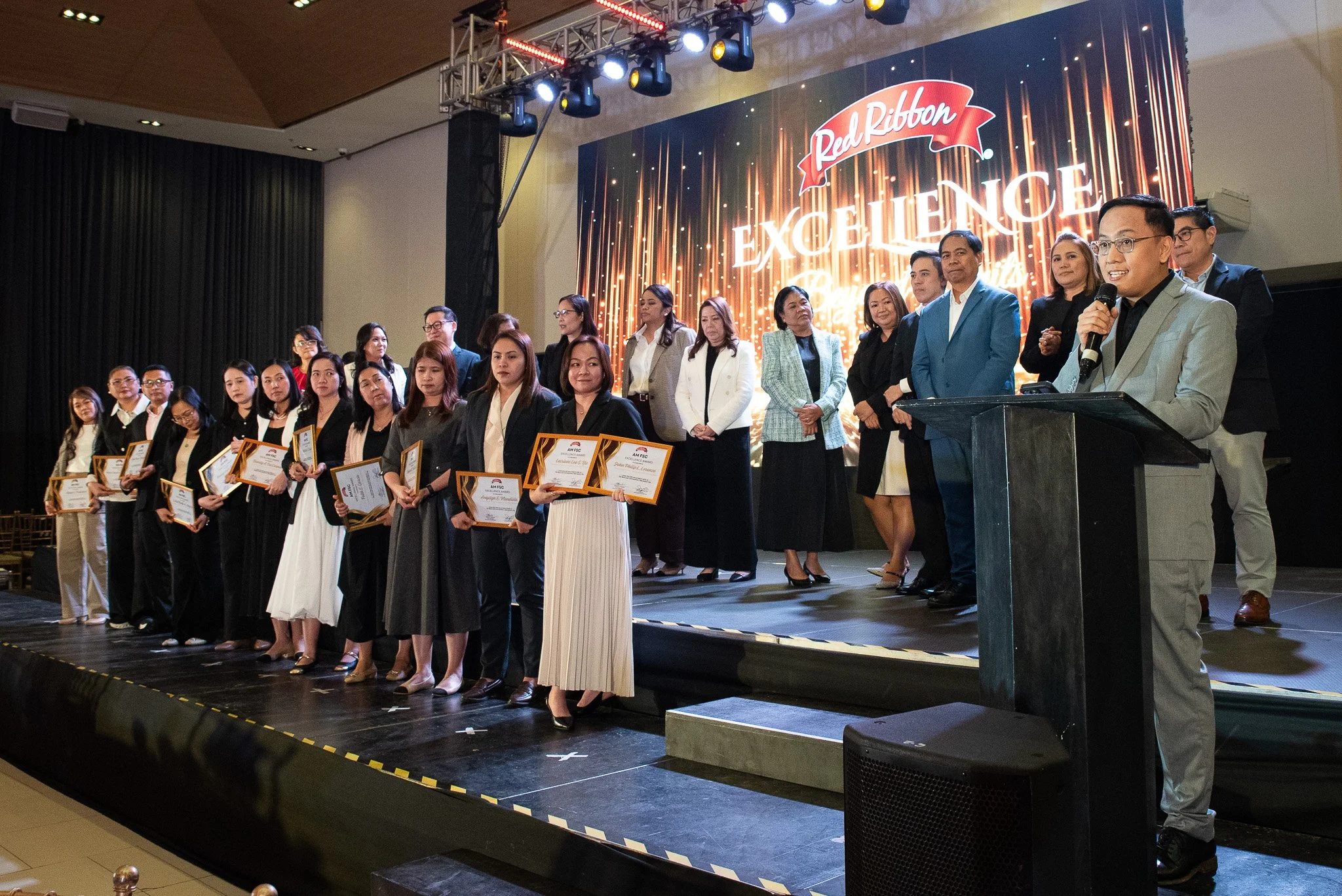 Group of people on stage at an award ceremony, with some holding certificates, under a large screen displaying 'Red Ribbon EXCELLENCE.'
