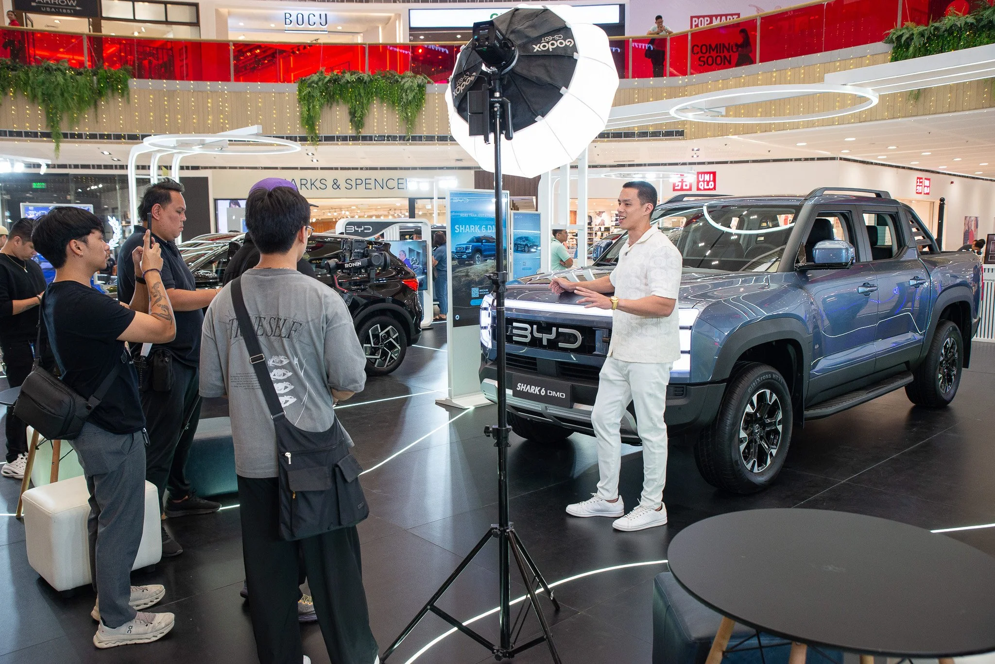 Man in white outfit filming a man in white shirt and pants standing next to a blue pickup truck with a camera setup and professional lighting at an indoor car exhibition.