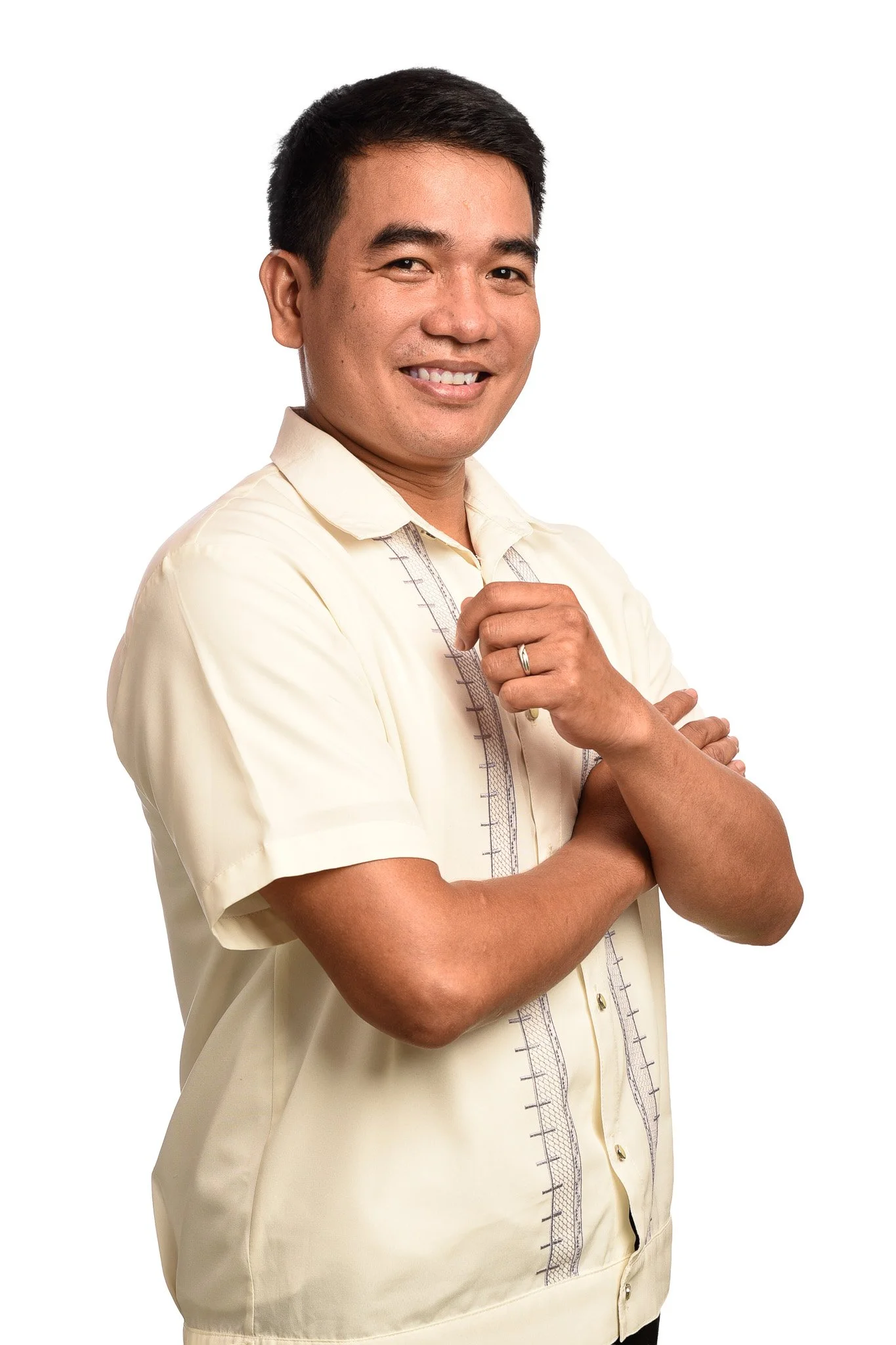 A smiling man with short black hair, wearing a beige Filipino Barong shirt, standing against a white background, with arms crossed and a confident pose.