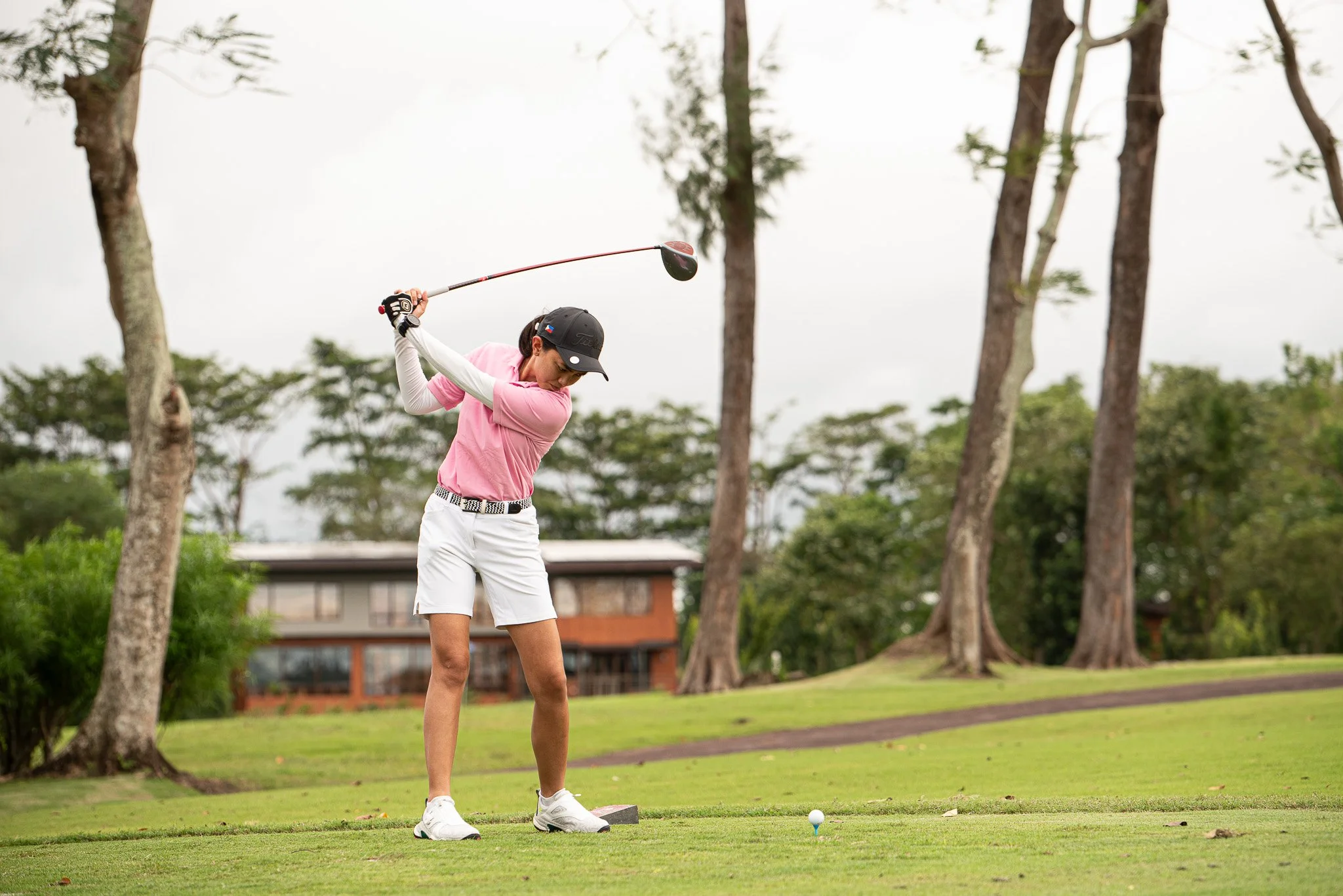 A woman in a pink shirt and white shorts preparing to hit a golf ball on a golf course with trees in the background.
