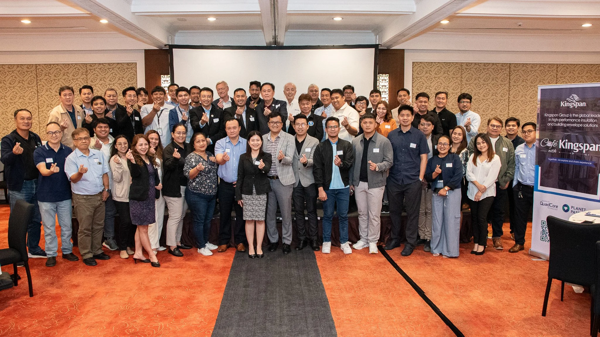 Group of people posing for a photo at a conference or seminar, standing on a stage and making a finger heart gesture, with a large screen and promotional banner in the background.