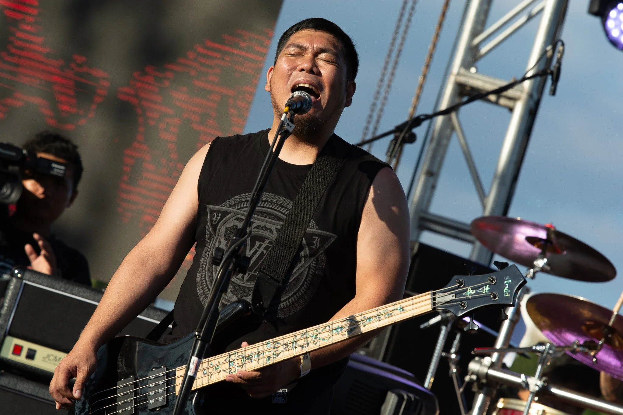 A male musician passionately singing into a microphone while playing a bass guitar during an outdoor concert, with stage equipment and drums visible in the background.