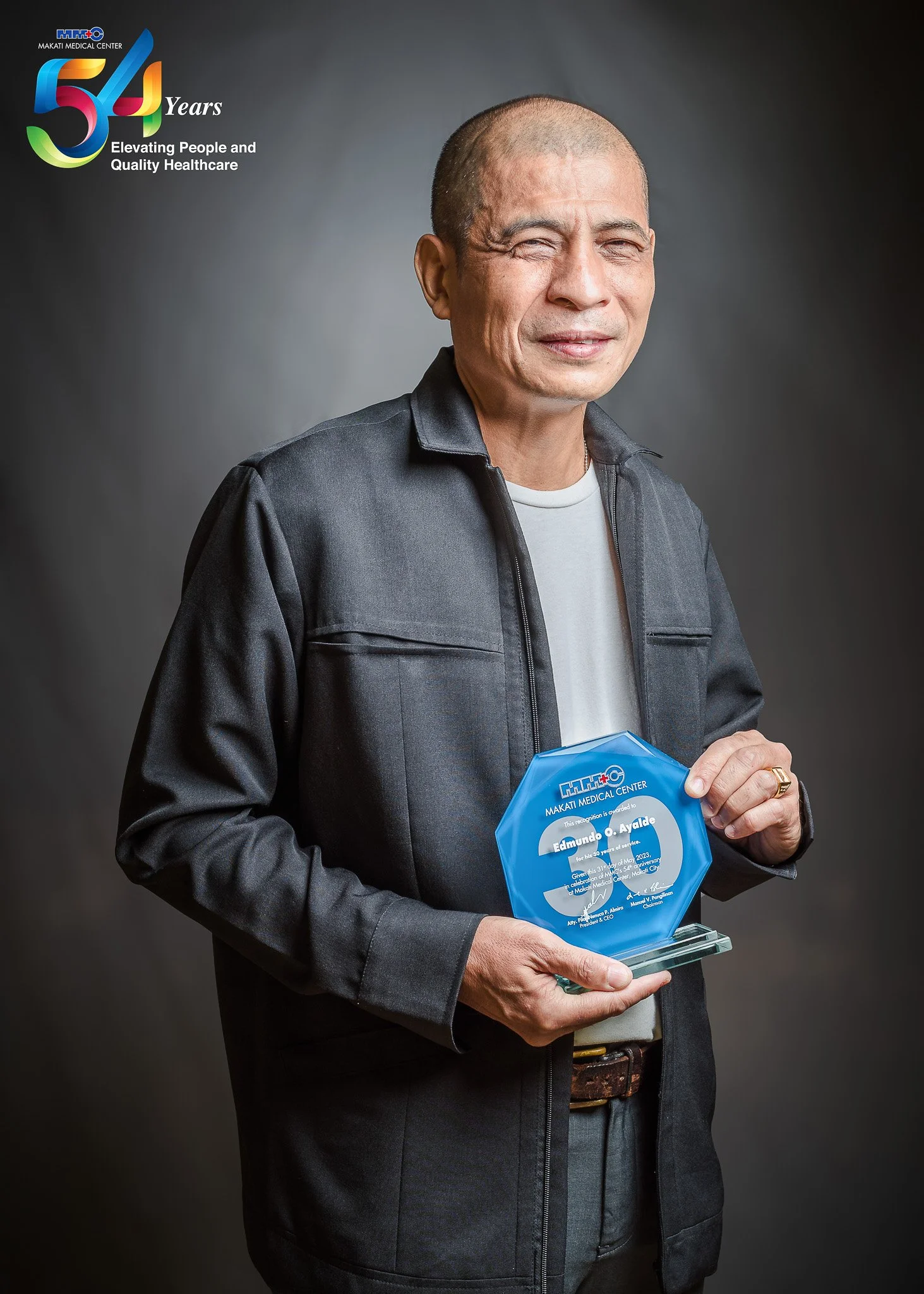 A man holding a commemorative plaque from Makati Medical Center celebrating 54 years. The man is wearing a black jacket and white shirt, standing against a dark background.