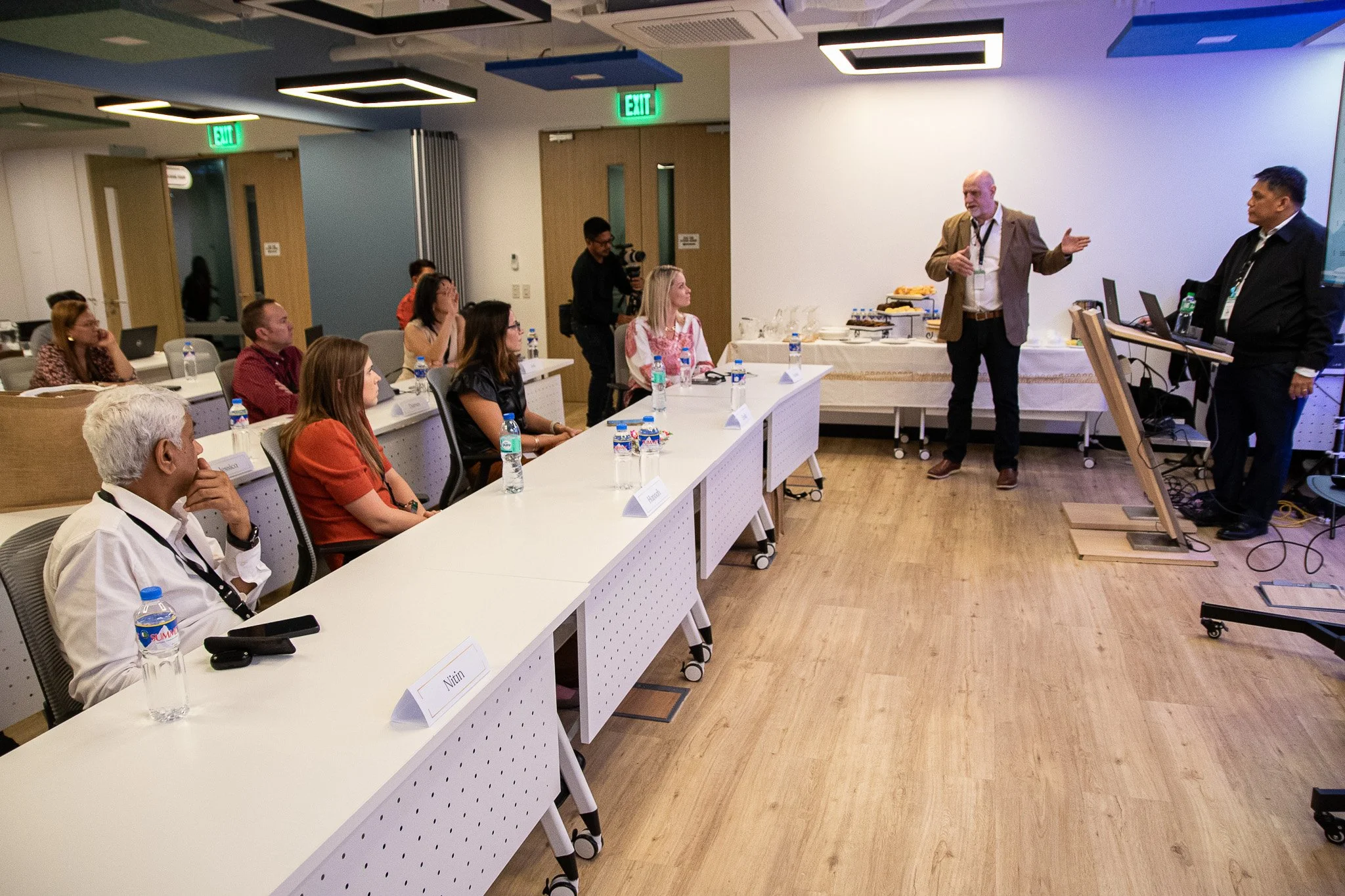 A group of people attending a presentation in a conference room. The speaker is standing near a podium, gesturing with his hands. Attendees are sitting at tables with water bottles, facing the speaker. There is a camera operator filming the event and