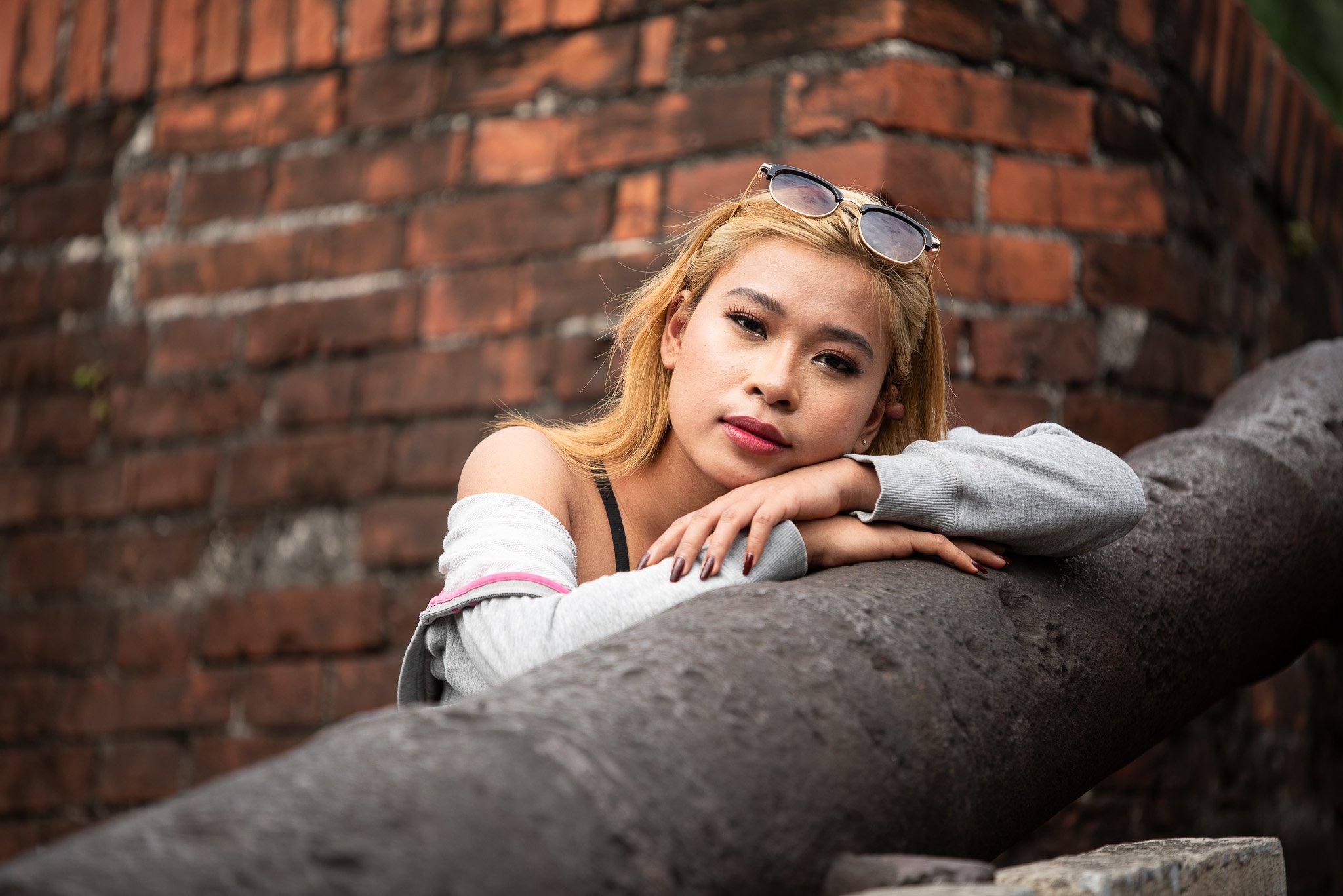 Young woman with blonde hair and sunglasses resting her head on her arms on a concrete pipe in front of a brick wall.