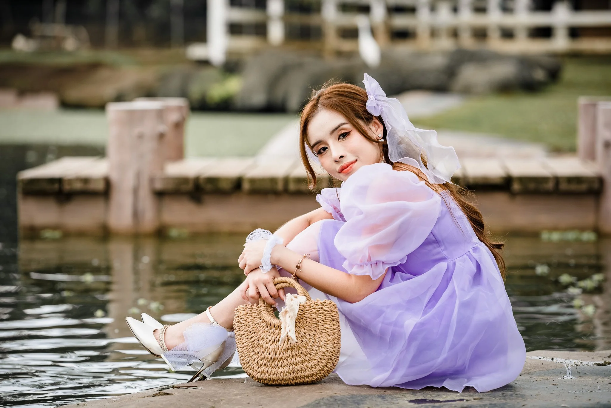 A young woman in a lavender dress sitting by a pond, holding her leg, with a woven handbag and white shoes, outdoors in a park-like setting.