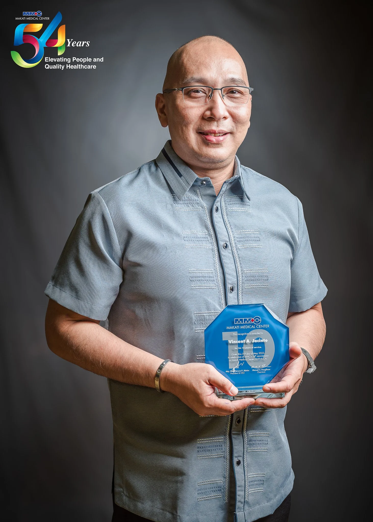 A man in a gray shirt holding a blue plaque from Makati Medical Center, celebrating 54 years of service.