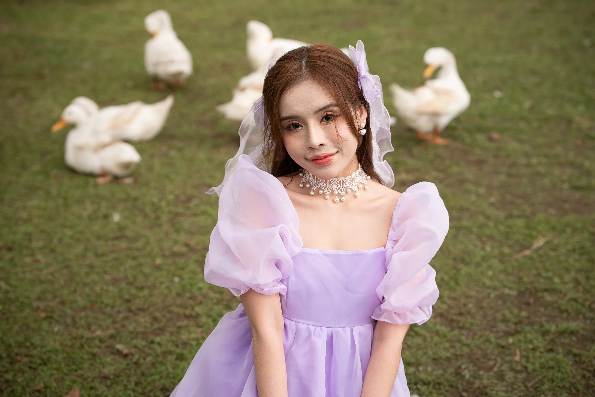 A woman in a lavender dress with puffed sleeves and a lace choker stands outdoors on grass, with several white ducks in the background.