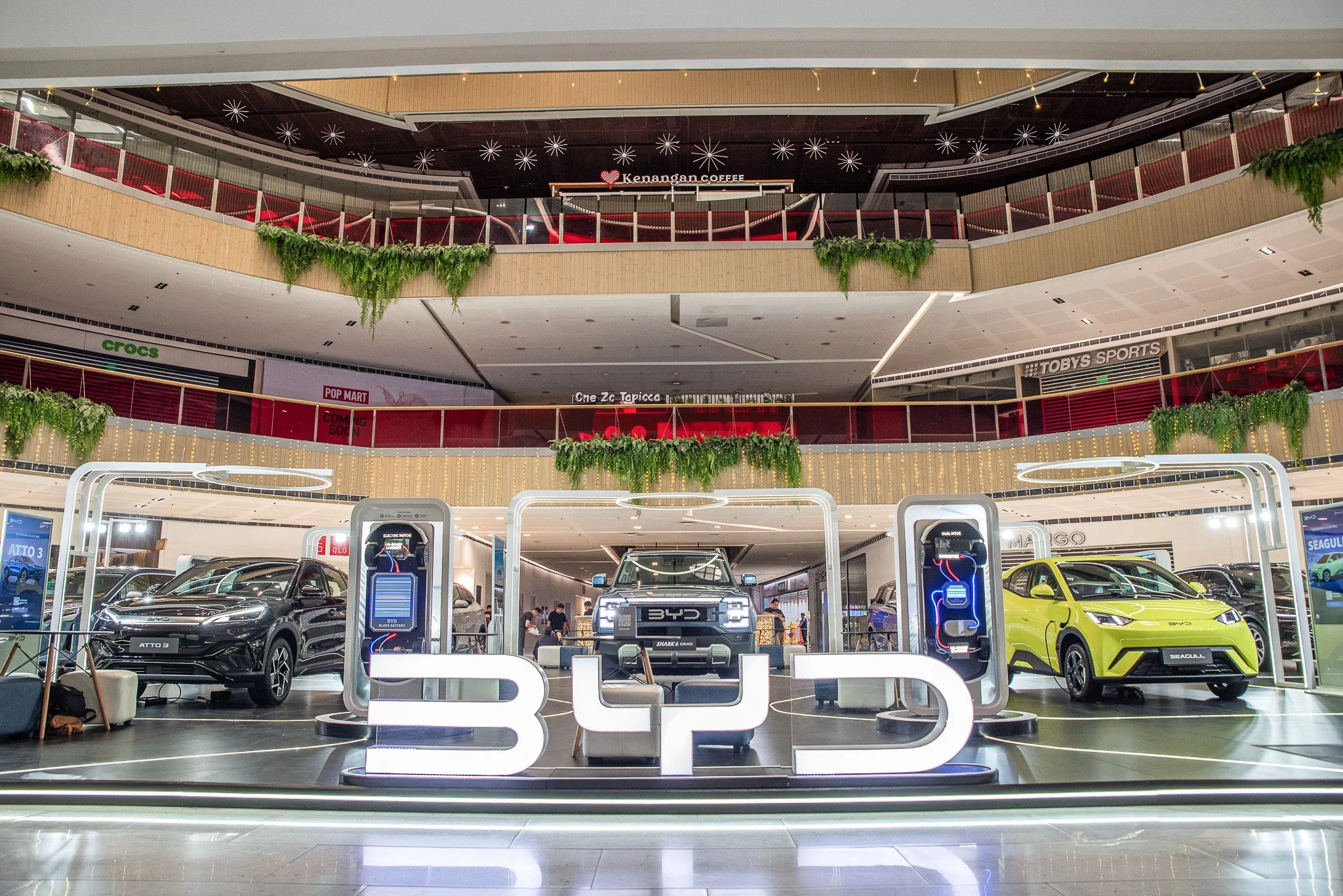 Display of three electric BYD cars in a shopping mall with neon sign reading 'BYD' and charging stations, upper floors with stores and hanging greenery.