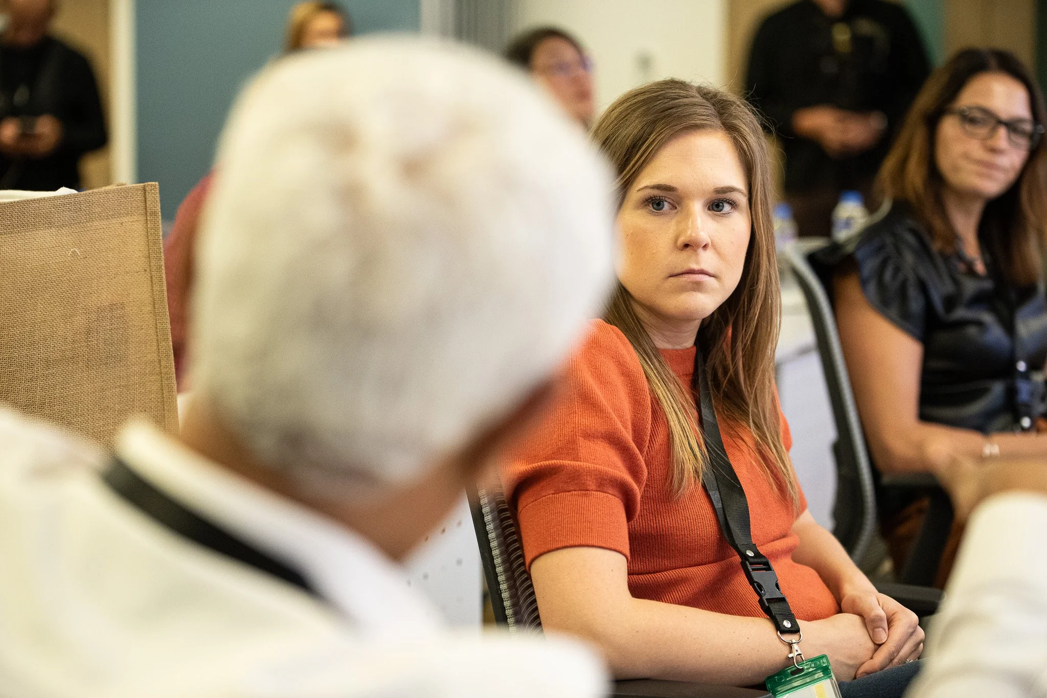 A woman with long brown hair and an orange shirt looking attentively at an elderly woman with white hair during a conference or meeting, with other seated women and a blurred background.