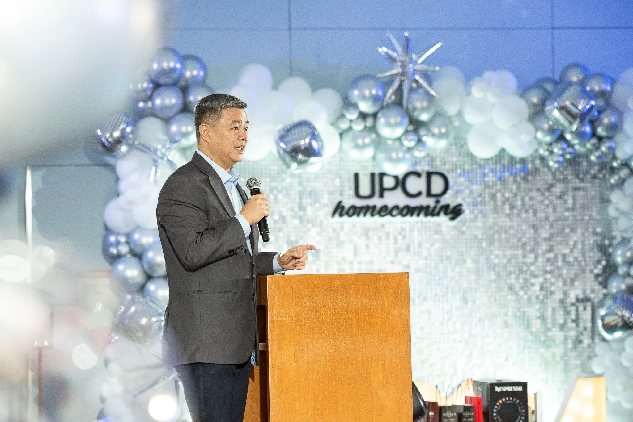 A man in a gray suit and light blue shirt holding a microphone and speaking at a podium during a UCSD homecoming event. The background decor includes silver balloons, white balloons, and a sign that reads 'UCSD homecoming.'