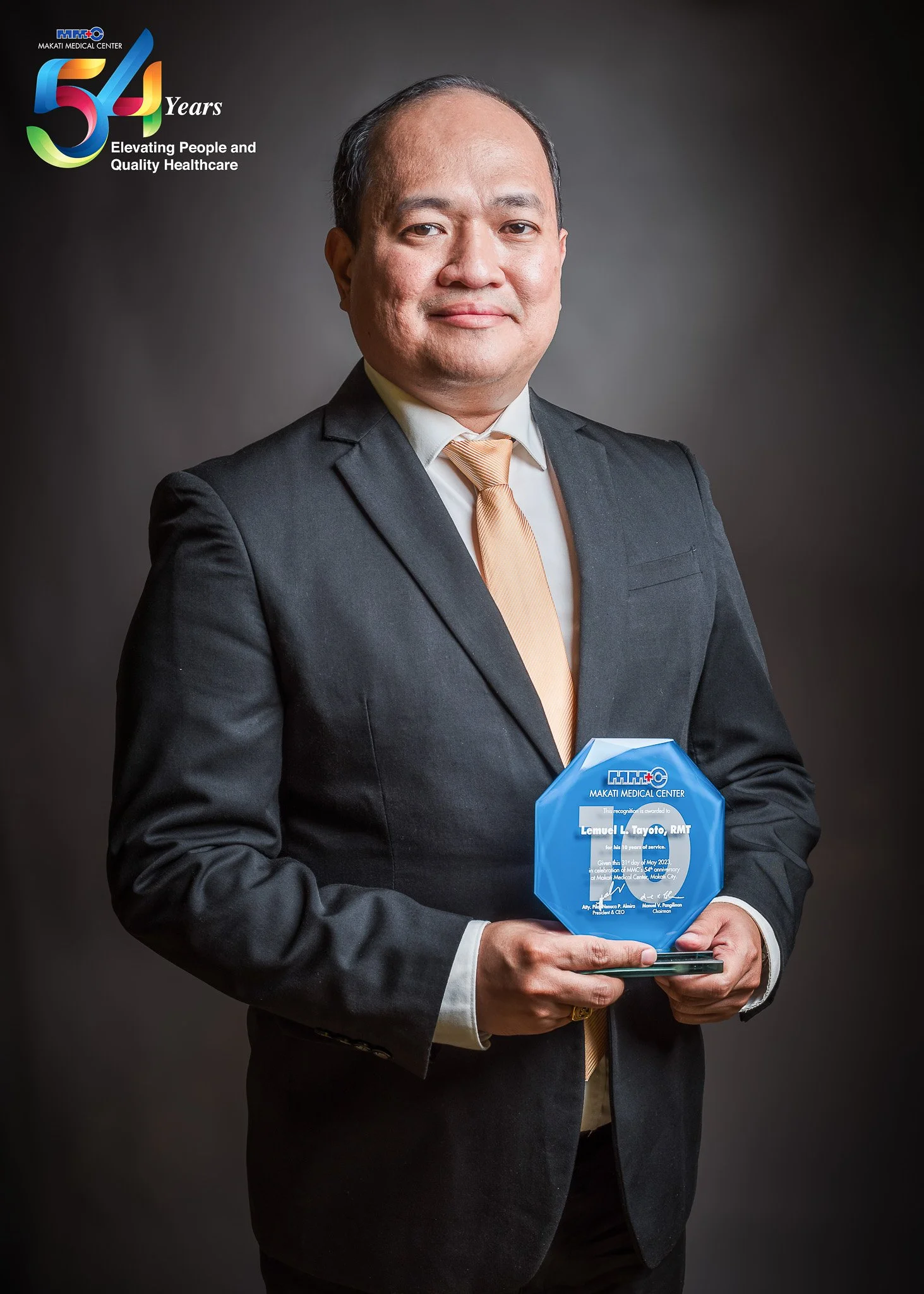 A man in a black suit and gold tie holding a blue award plaque with the Makati Medical Center logo on it. The background is dark. The top left corner has the Makati Medical Center 54 years anniversary logo.
