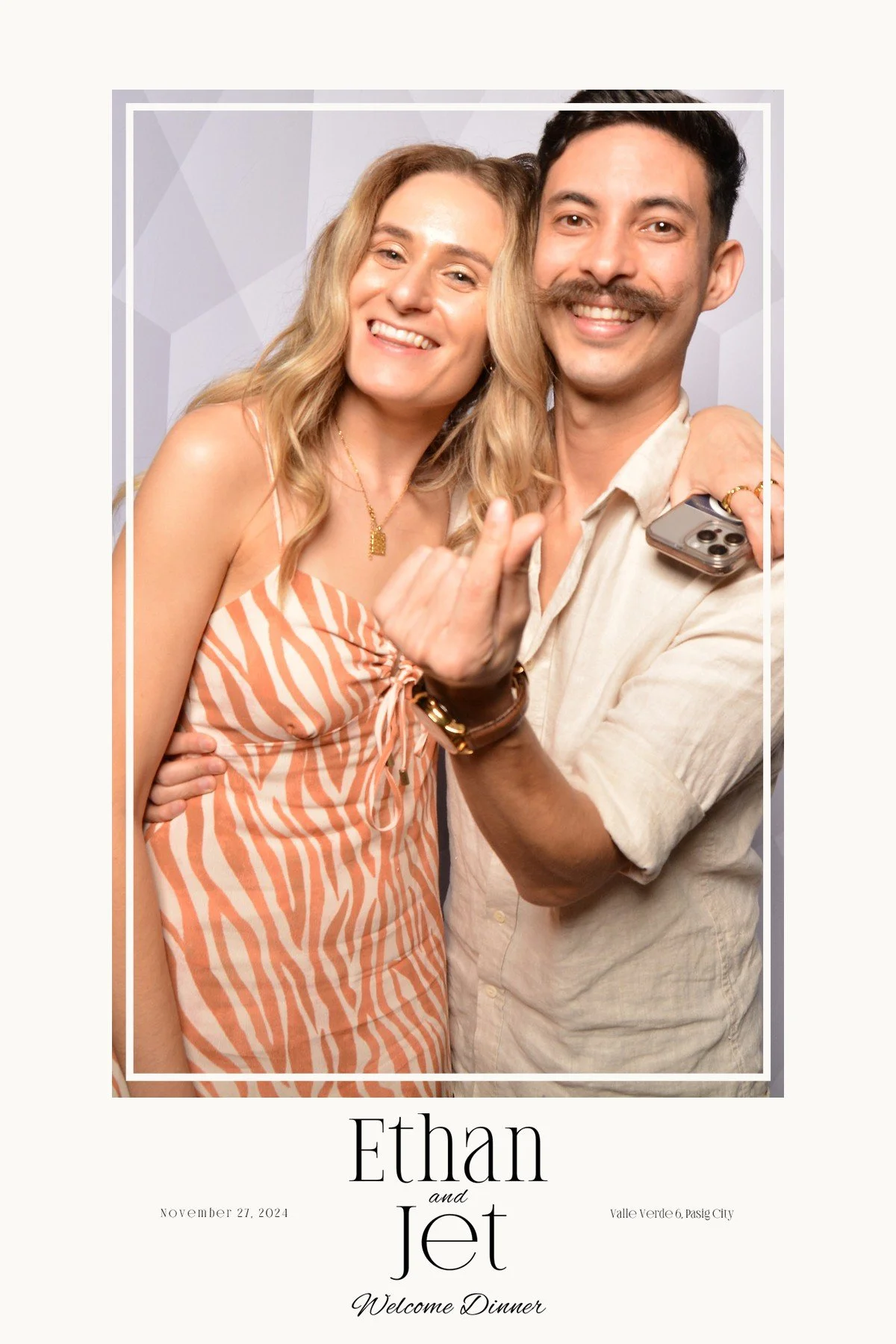 A smiling couple at a wedding or event, standing close together with one arm around each other. The woman has blonde hair and is wearing a striped dress, and the man has dark hair and a mustache, wearing a light-colored shirt. The man is holding a sm