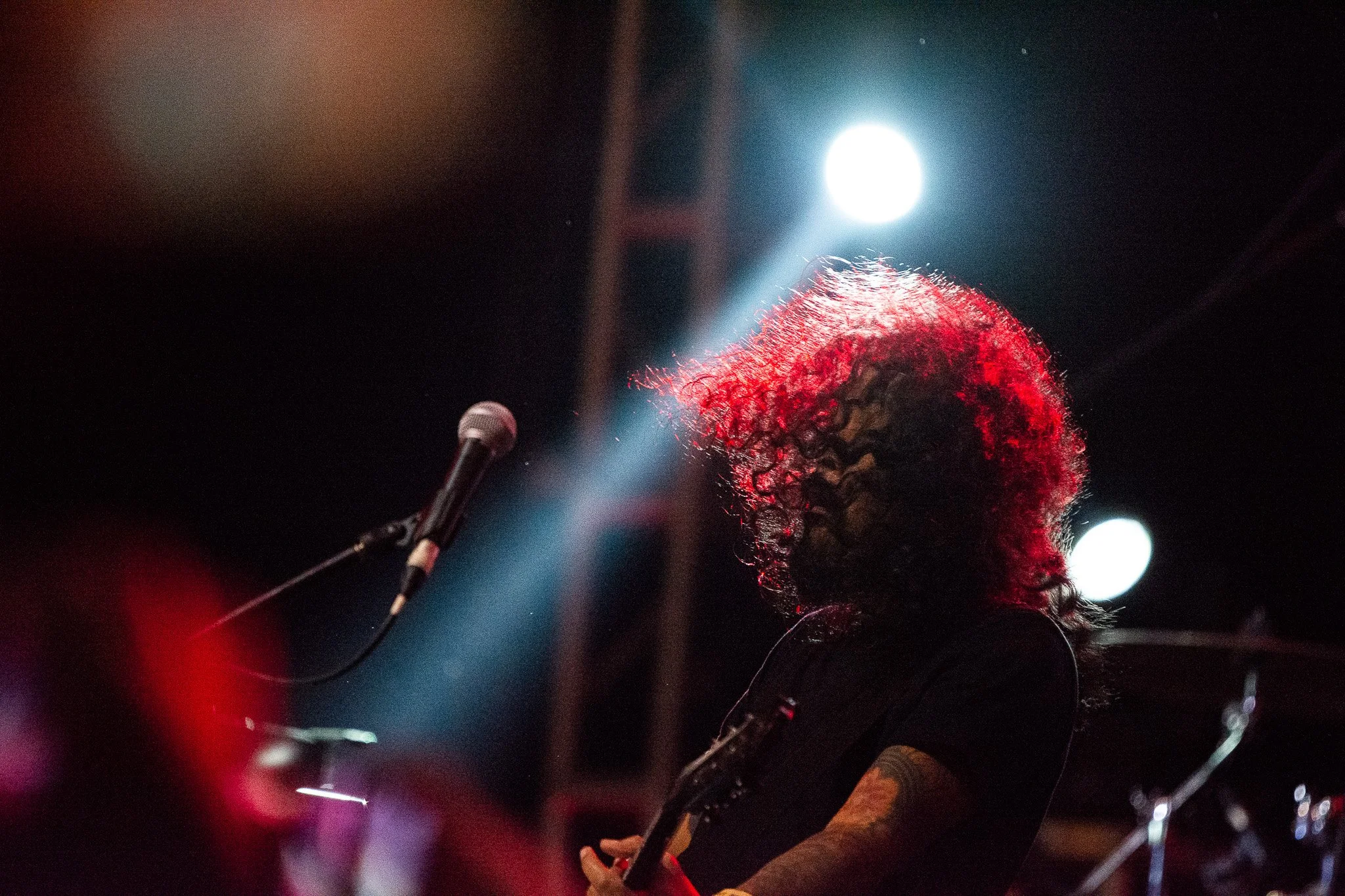 A musician with curly hair illuminated by stage lights, performing on stage with a microphone in front of her.