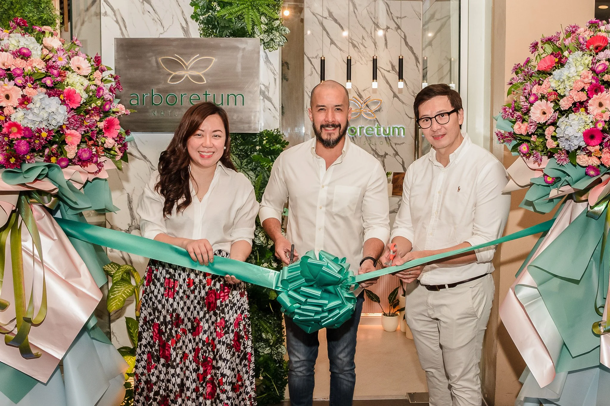 Three people at a ribbon-cutting ceremony in front of a floral arrangement and a sign that says 'arboratum'.