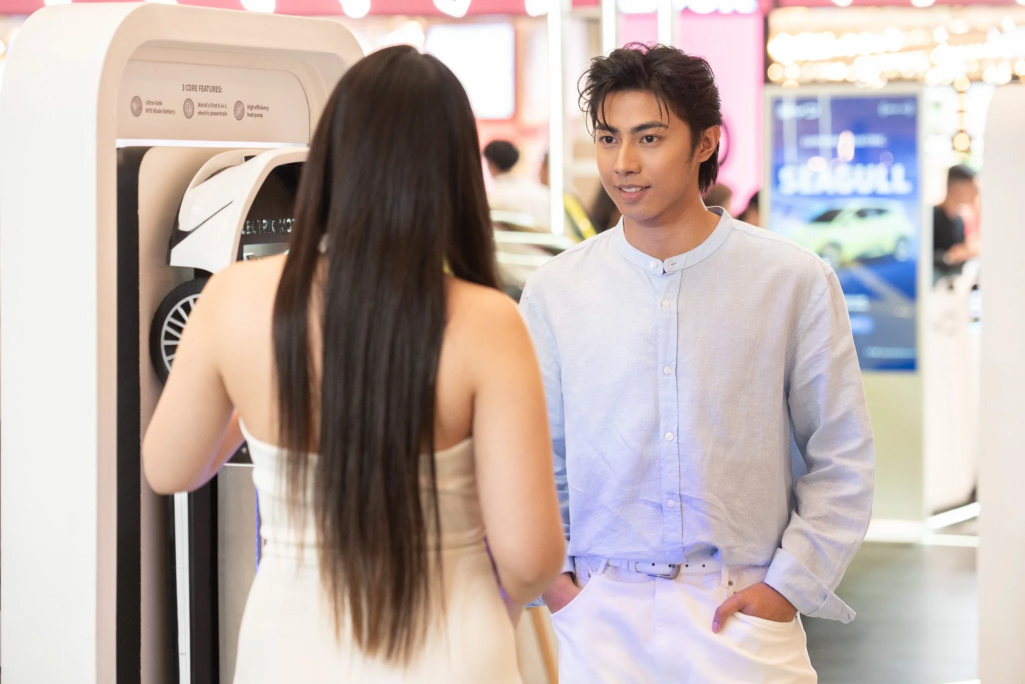A young man and woman with long dark hair are having a conversation at an electric vehicle charging station inside a shopping mall or indoor space. The man is dressed in a light blue shirt and white pants, and the woman is wearing a sleeveless beige 