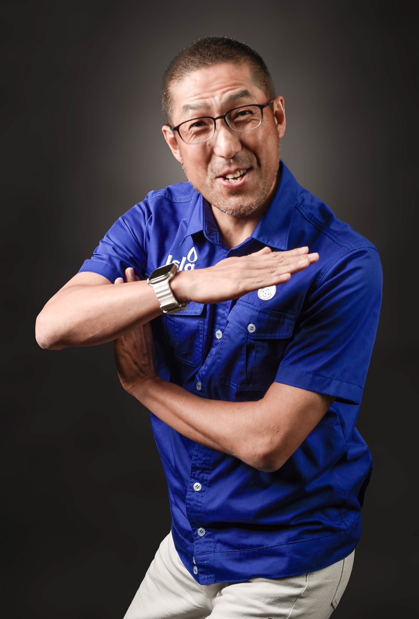 A man in a blue collared shirt making a gesture with his hand across his chest, smiling and winking at the camera, wearing glasses and a watch, against a dark background.
