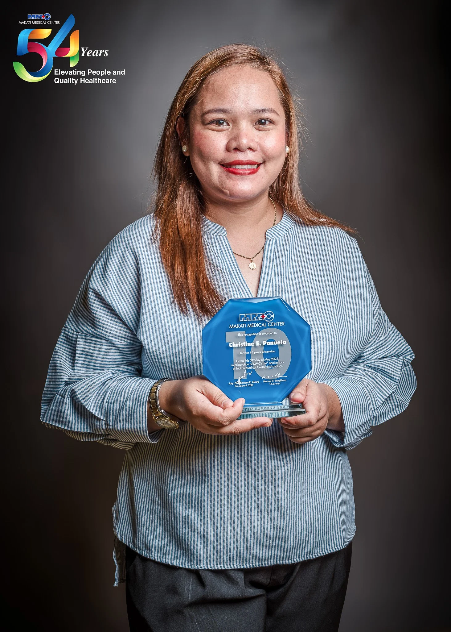 Woman holding an award plaque with a dark background, wearing a light blue striped blouse, smiling, with a logo and celebrating 54 years of Makati Medical Center.