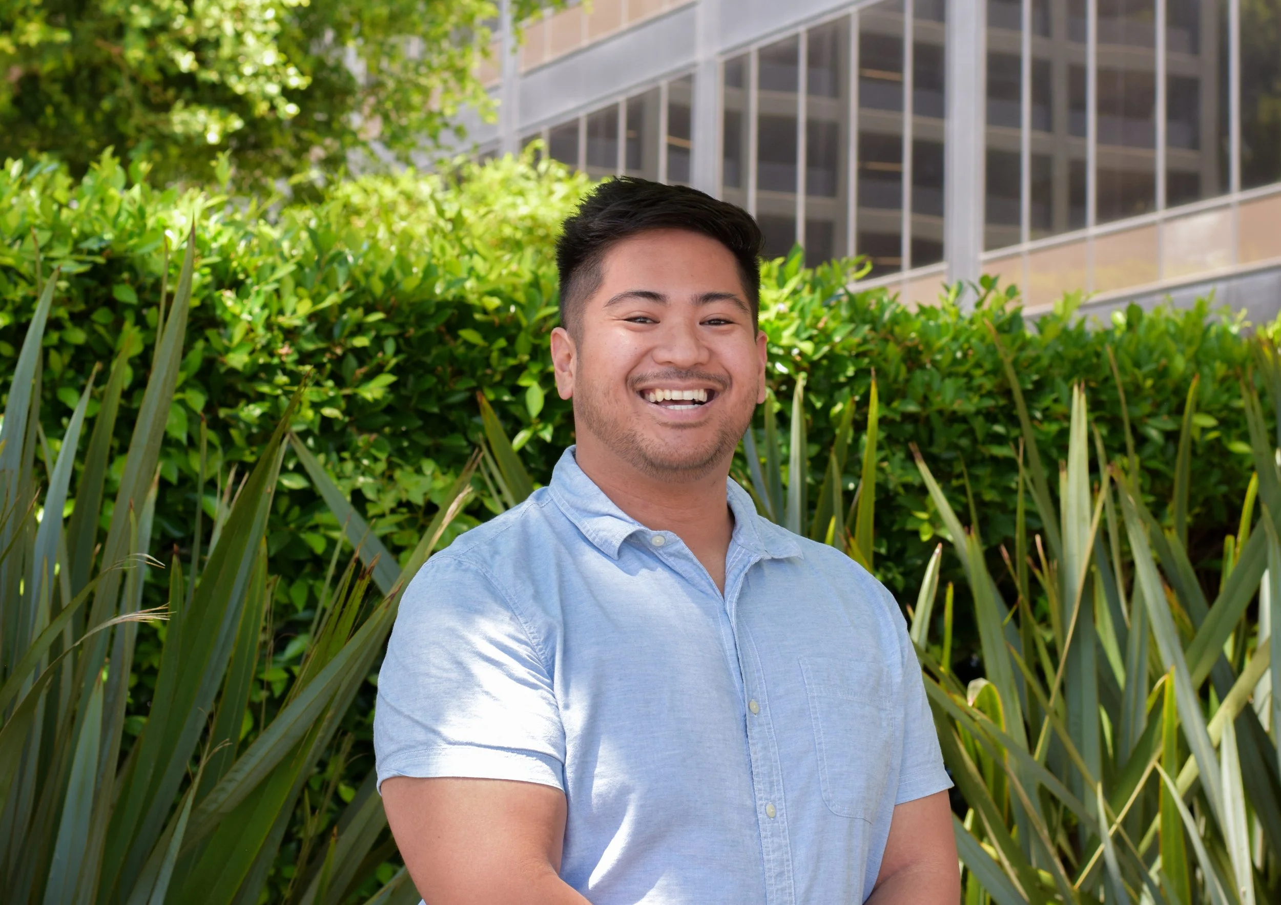 A happy young man with short black hair and a light blue button-up shirt, smiling outdoors in front of green bushes and a building with glass windows.