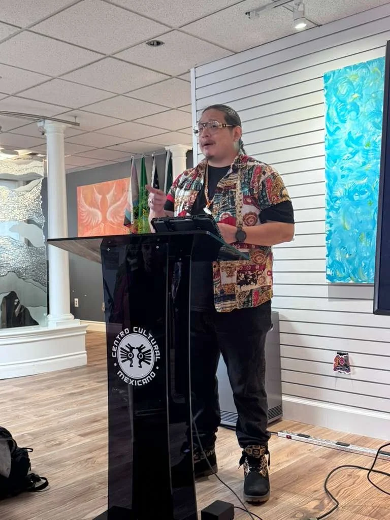 A man with glasses and long hair tied back, wearing a colorful patterned shirt over a black T-shirt, standing at a podium labeled 'Centro Cultural Mexicano'. He is speaking and gesturing with his right hand, with artwork on the wall behind him.