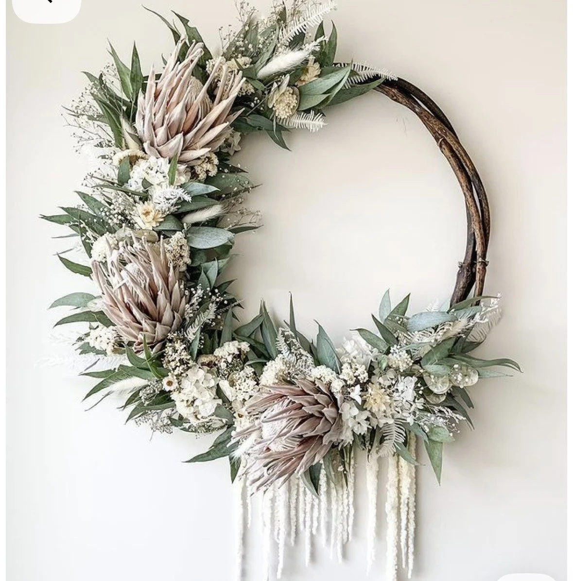 Decorative wreath with large protea flowers, eucalyptus leaves, and white hanging beads on a plain wall.