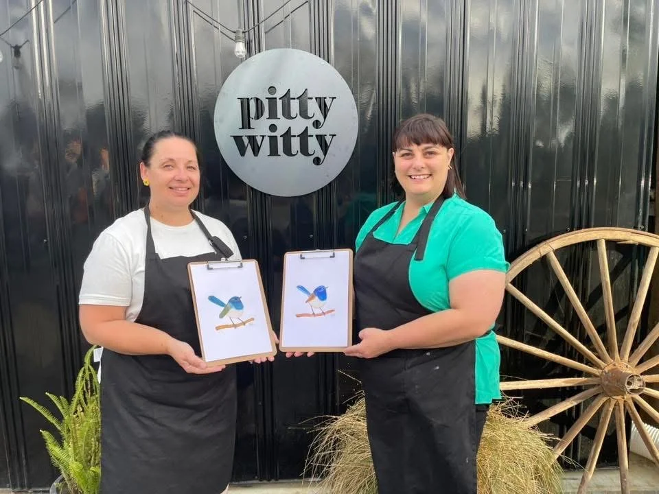 Two women wearing black aprons holding clipboards with a picture of a bird on it, standing outside a building with a sign that says 'pity witty', a hay bale, a wooden wagon wheel, and some plants nearby.
