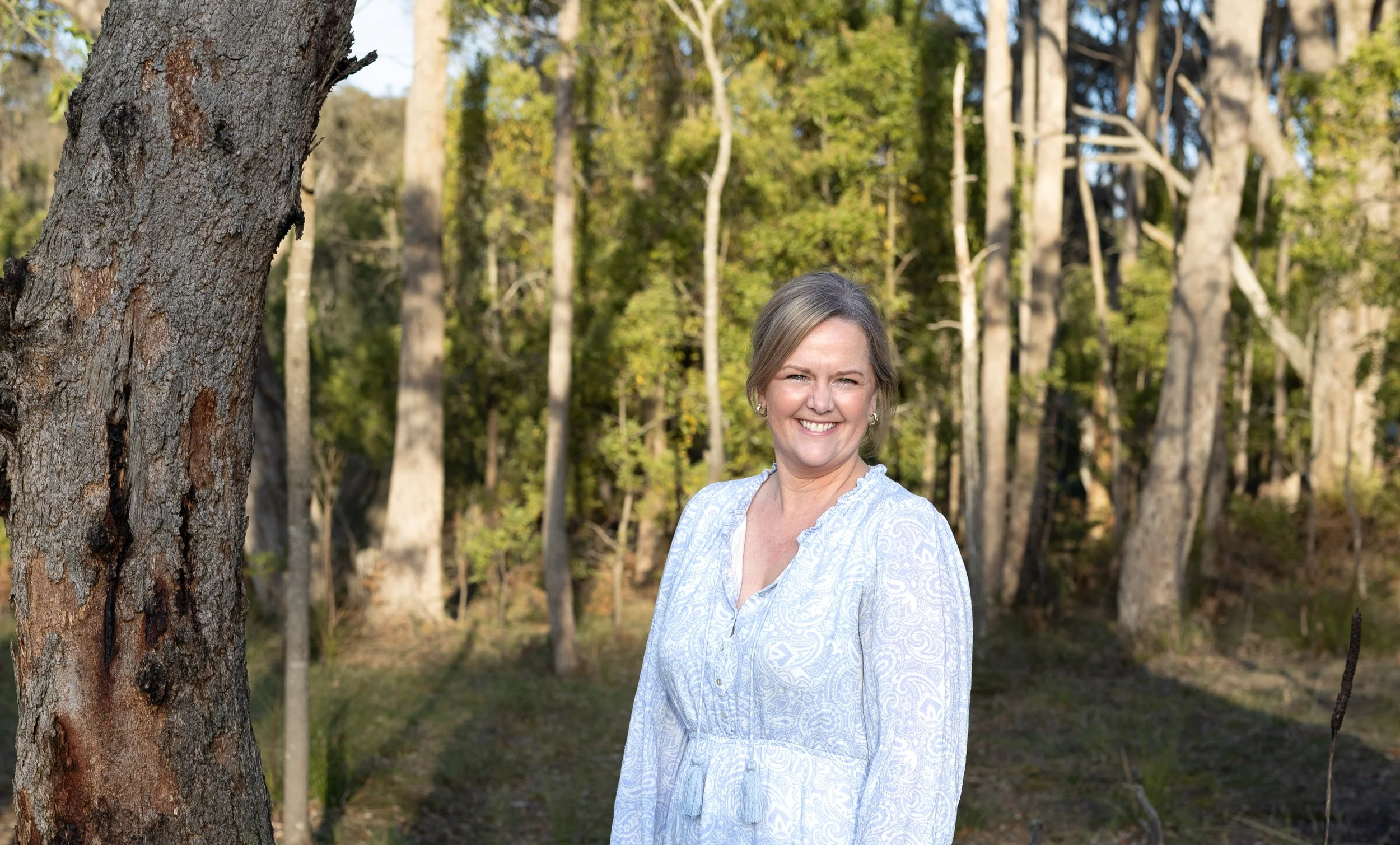A smiling woman with short blonde hair wearing a white patterned blouse, standing in a forest during daylight, with trees and greenery in the background.