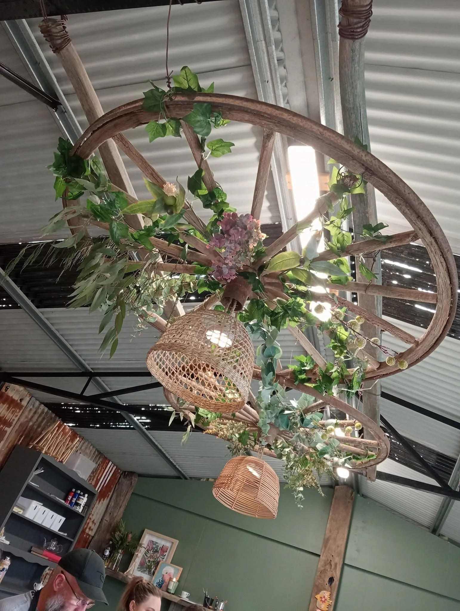 Decorative ceiling fixture made from an old wagon wheel adorned with hanging greenery, flowers, and woven lampshades.