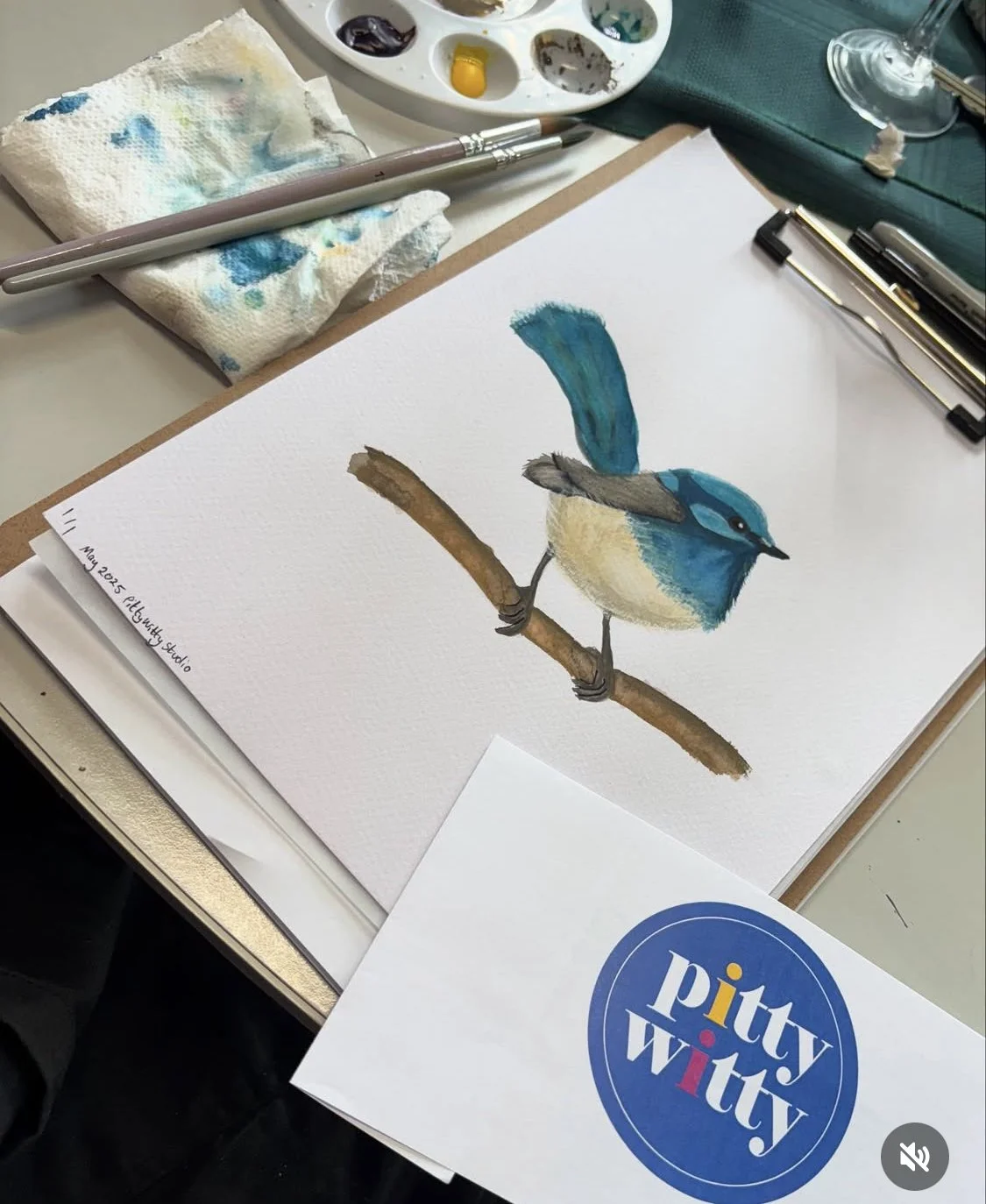 Watercolor painting of a blue bird perched on a branch, with painting supplies and a Pitt Artist Pen box in the background.