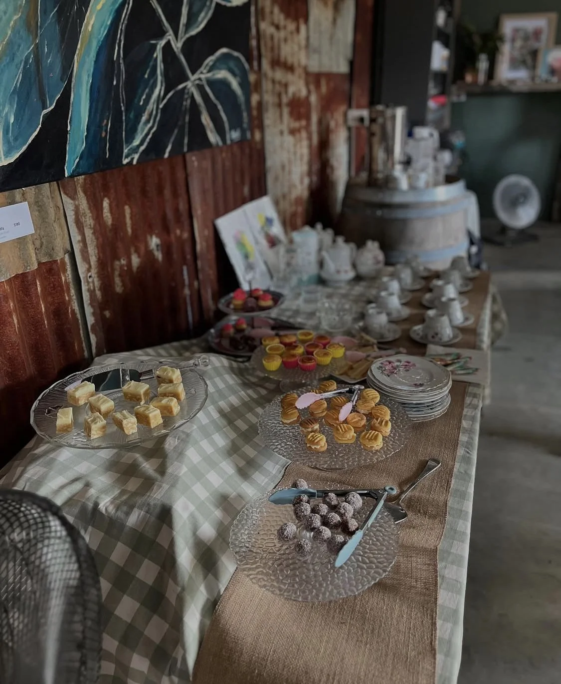 A dessert table with various sweet treats including blondies, cupcakes, truffles, and other confections arranged on glass and ceramic plates on a checkered tablecloth with a burlap runner, against a rustic wooden and metal background with artwork and