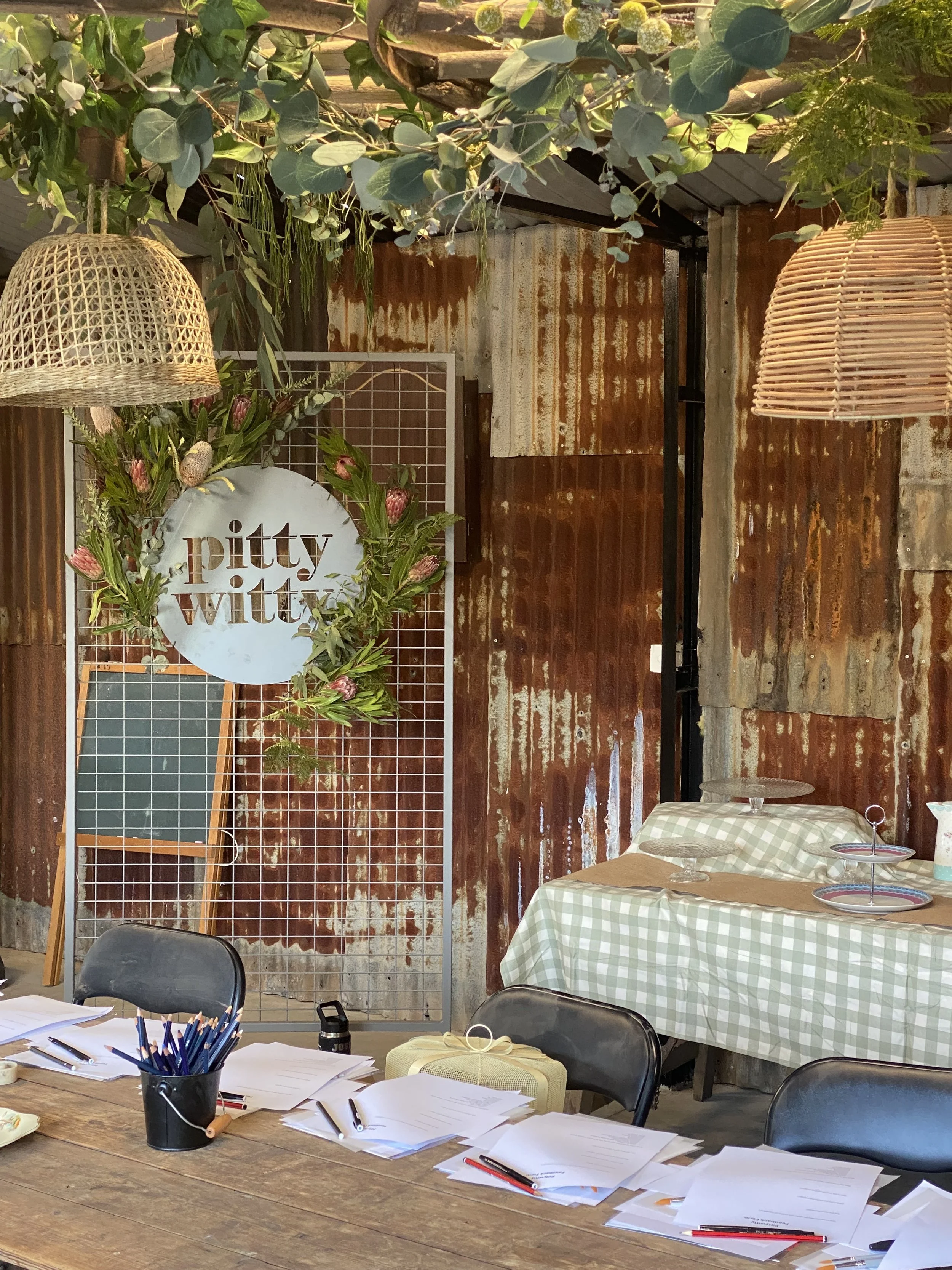 Rustic indoor space decorated with hanging wicker lamps, a floral wreath with the words "pitty witty" on a circular sign, a white board, a table with papers and pens, and a table with a checkered tablecloth and empty cake stands