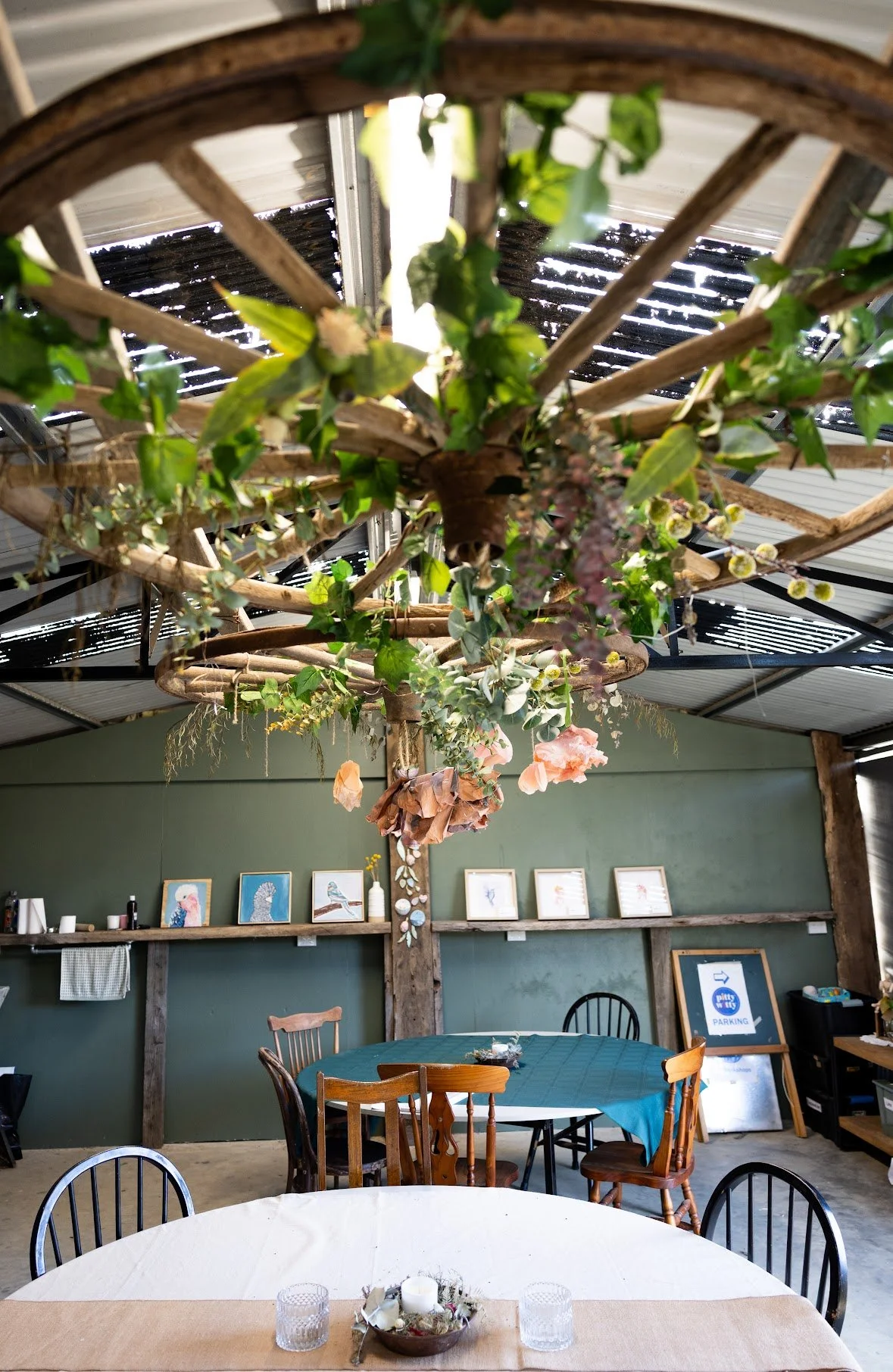 Interior of a rustic dining space with wooden beams, green walls, and framed artwork. Hanging from the ceiling is a decorative wooden wheel with greenery and flowers. There are tables with tablecloths and chairs around, and a sign in the background i