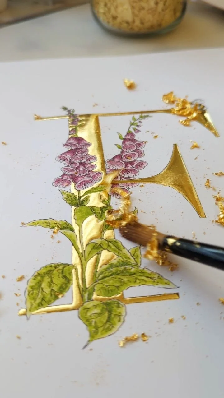 Hand-painted floral illustration with gold accents on white paper, with a brush applying gold paint and a bowl of rice in the background.