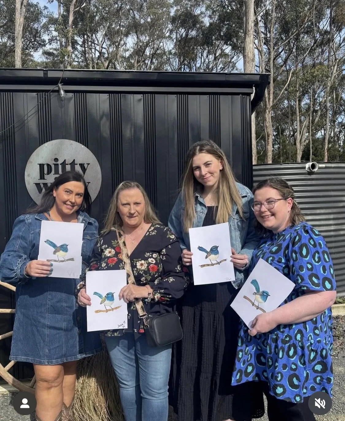 Four women standing outside in front of a black wall with a round sign that reads 'pitty' with the yellow word underneath, holding up drawings of a blue bird on a branch.