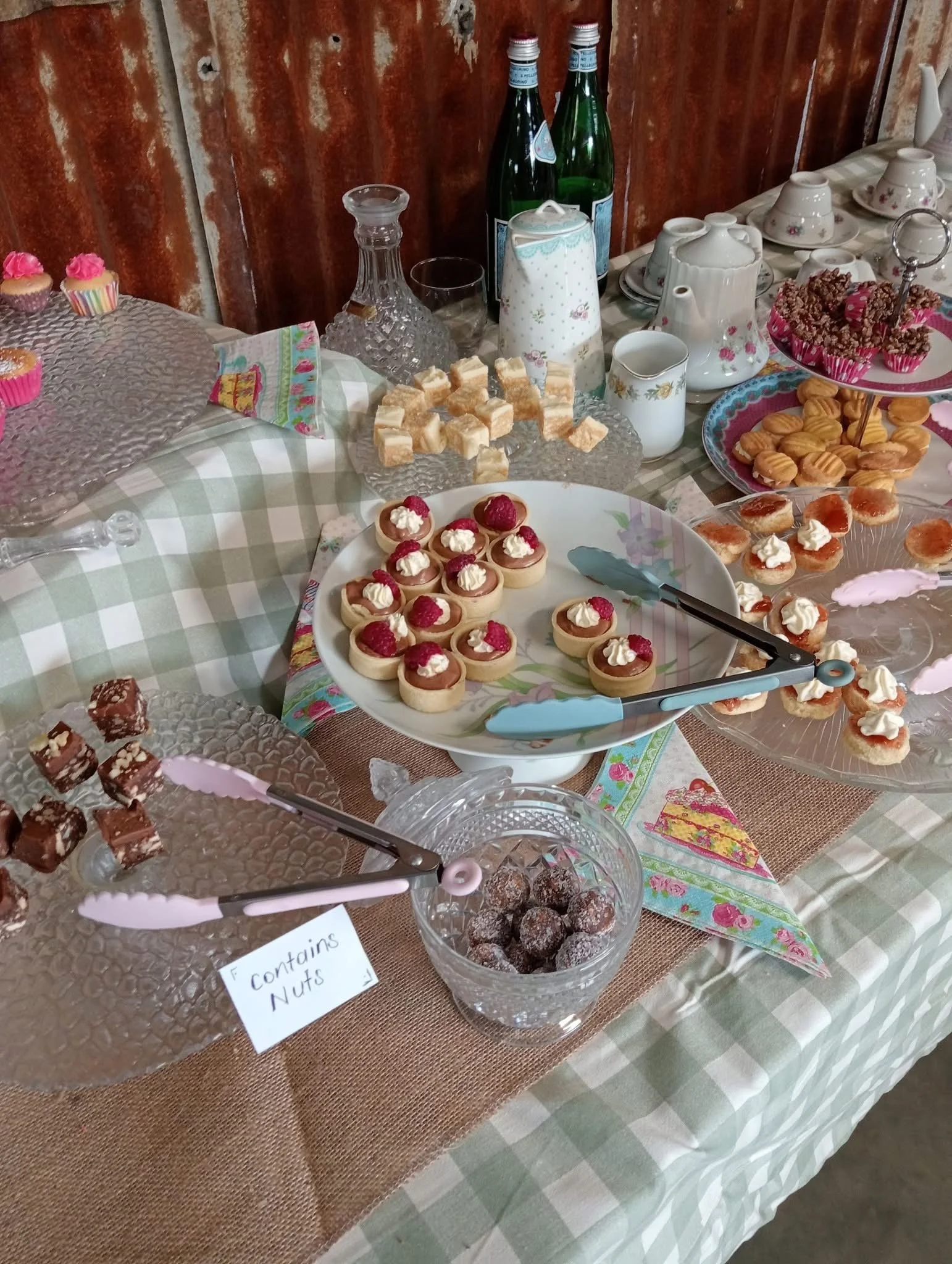 A table with an assortment of desserts, including raspberry-topped tarts, chocolate treats labeled "fountains nuts," and other sweets, with dishes, cups, bottles, and a rustic wooden wall in the background