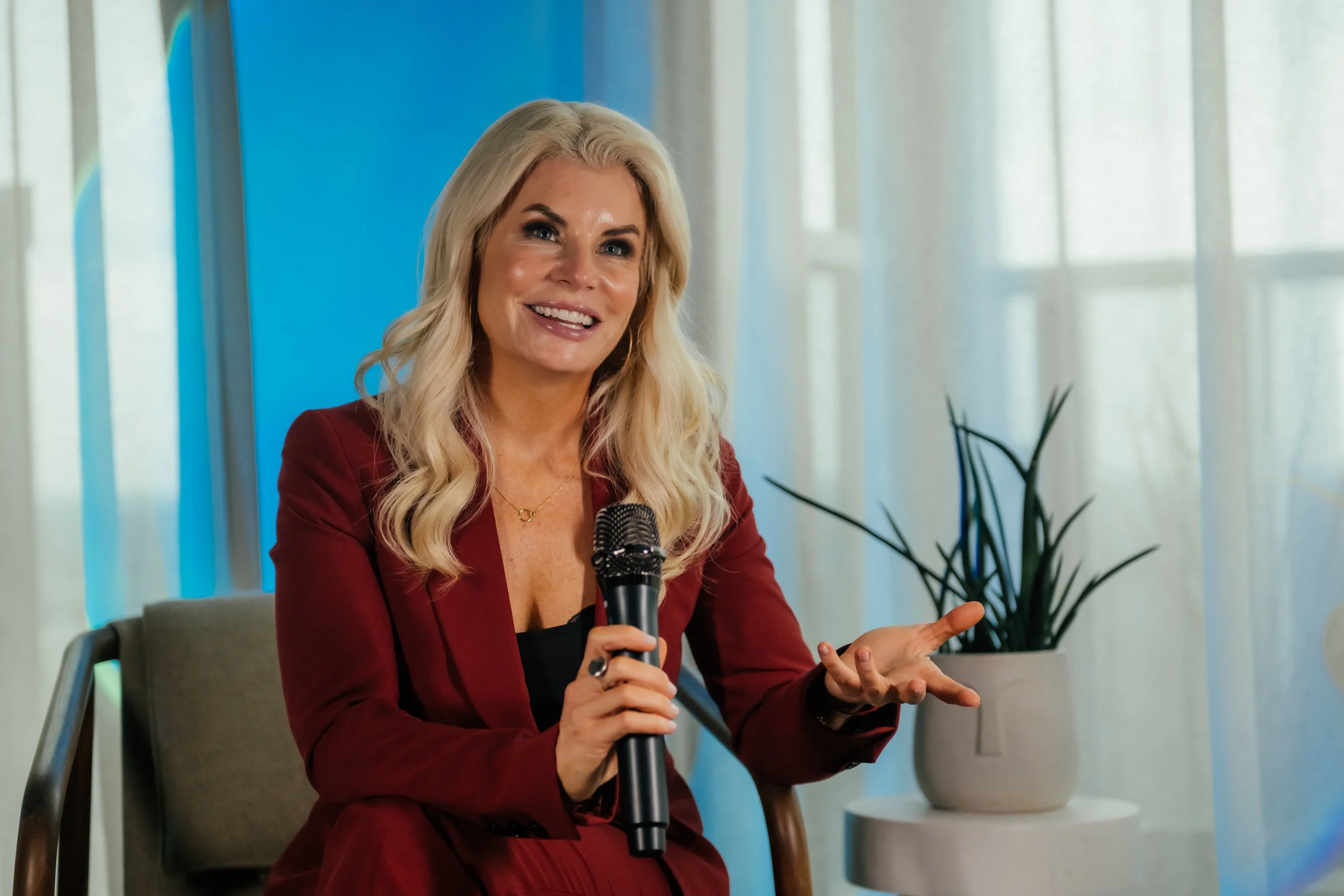 A woman with long blonde hair, wearing a red blazer, sitting on a chair, smiling, holding a microphone and gesturing with her hand.