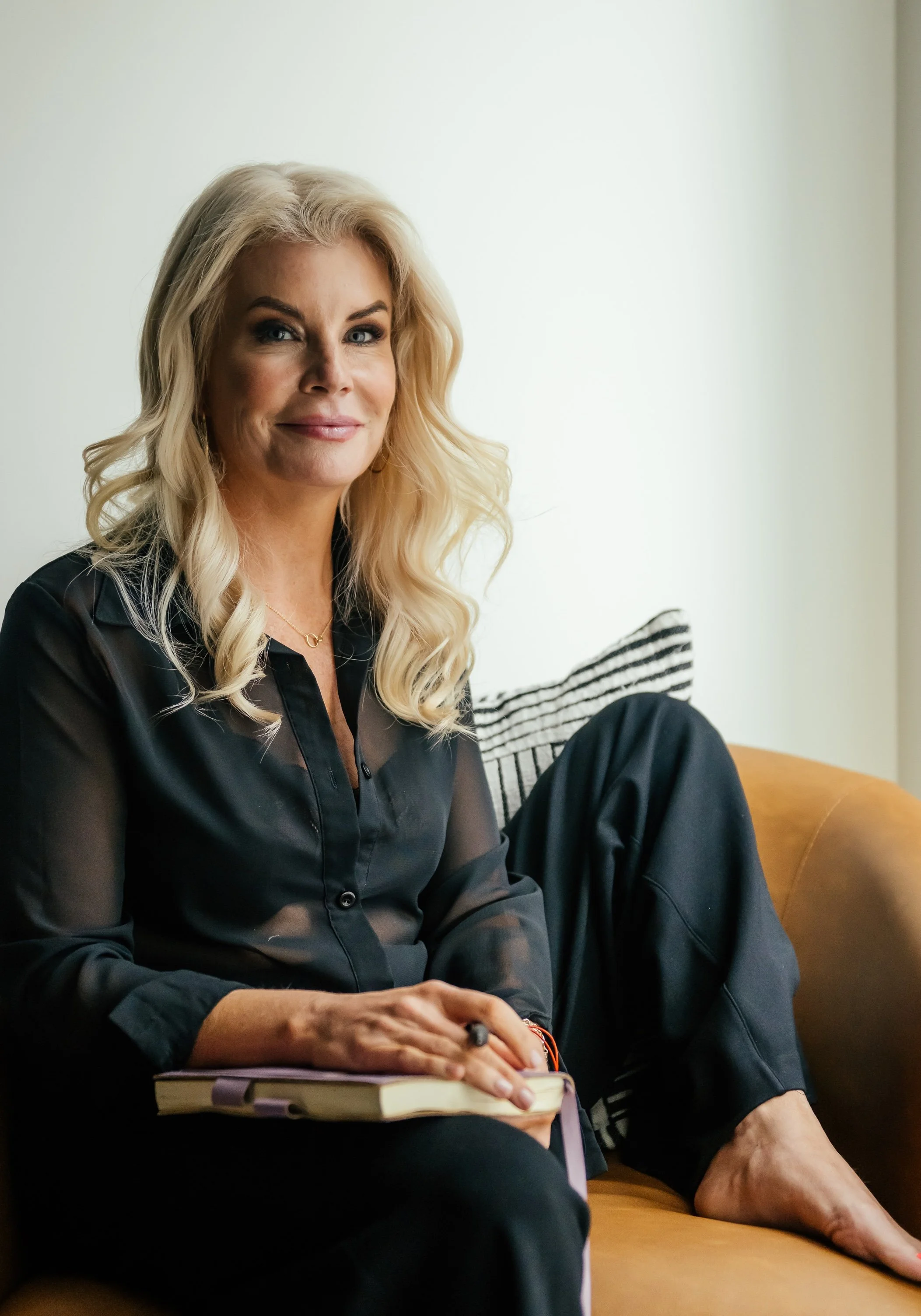 A woman with long blonde hair sitting on a tan couch, wearing a black sheer blouse and black pants, holding a book in her lap, and smiling at the camera.