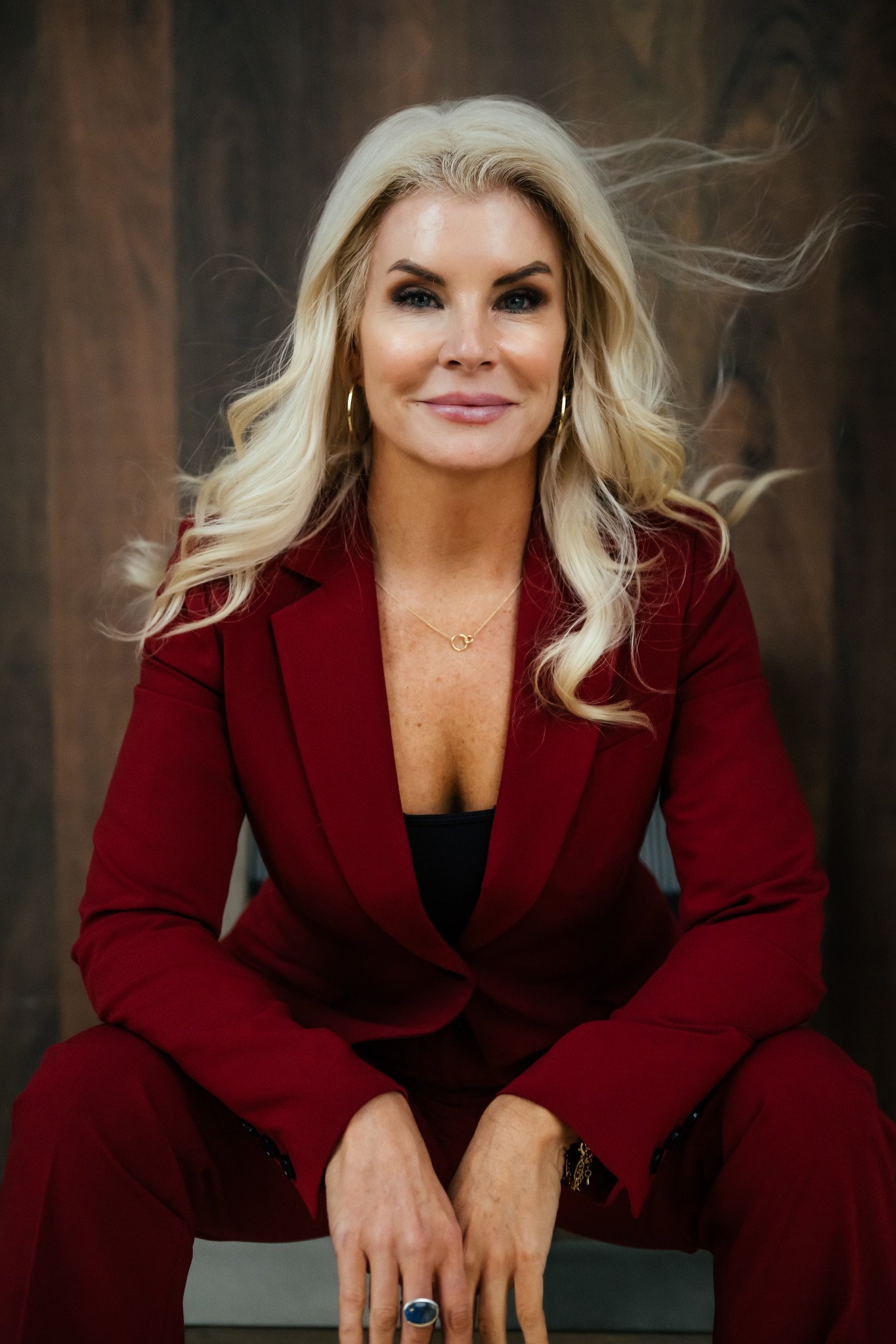 A woman with long blonde hair wearing a red blazer and matching pants, sitting and smiling at the camera against a dark wooden background.