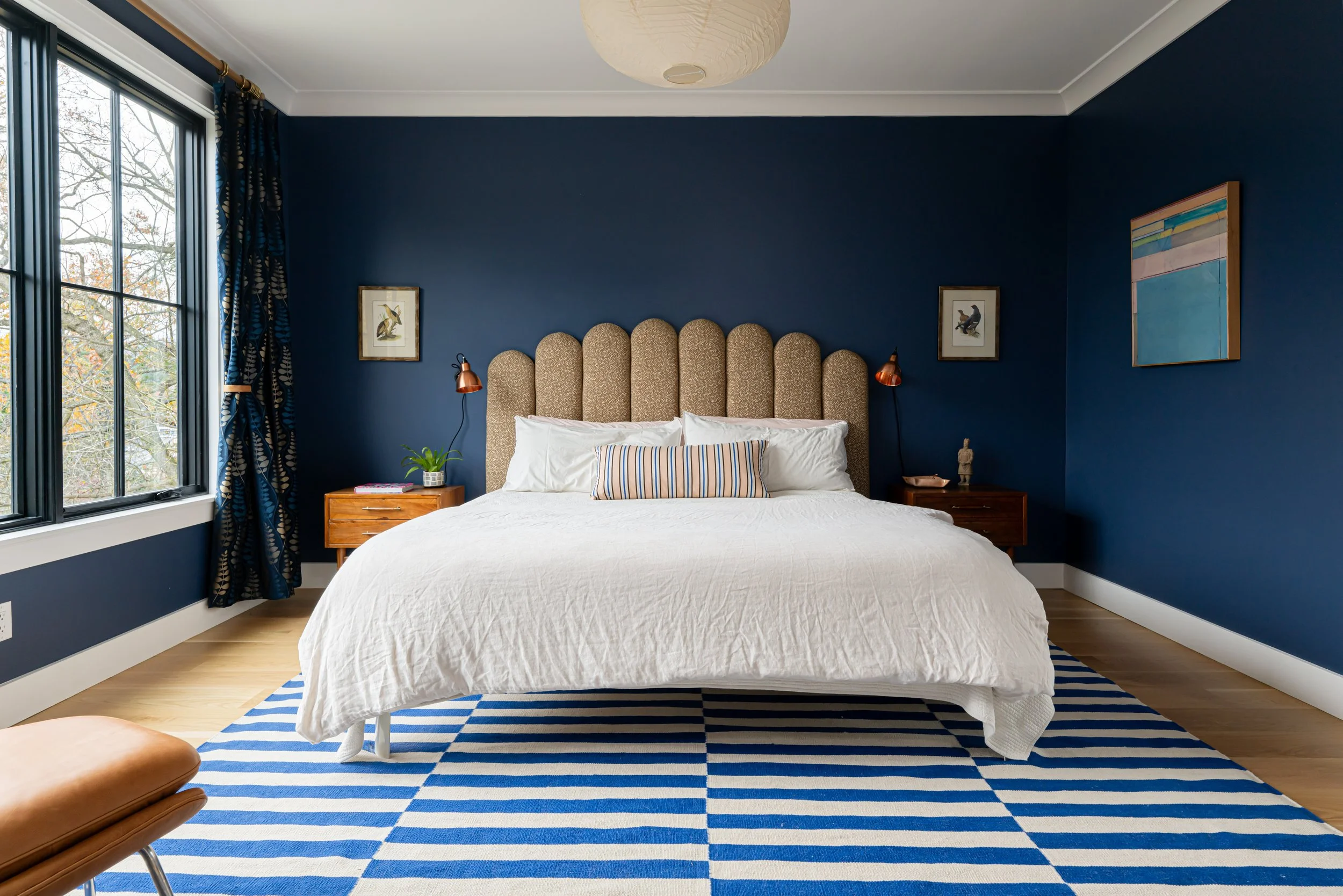 A bedroom with dark blue walls, a beige upholstered headboard, white bedding, and a striped pillow. There are two wooden nightstands with small lamps, framed artwork on the walls, and a large window with dark curtains. A blue and white striped rug is partially visible under the bed, and a leather chair is in the foreground.