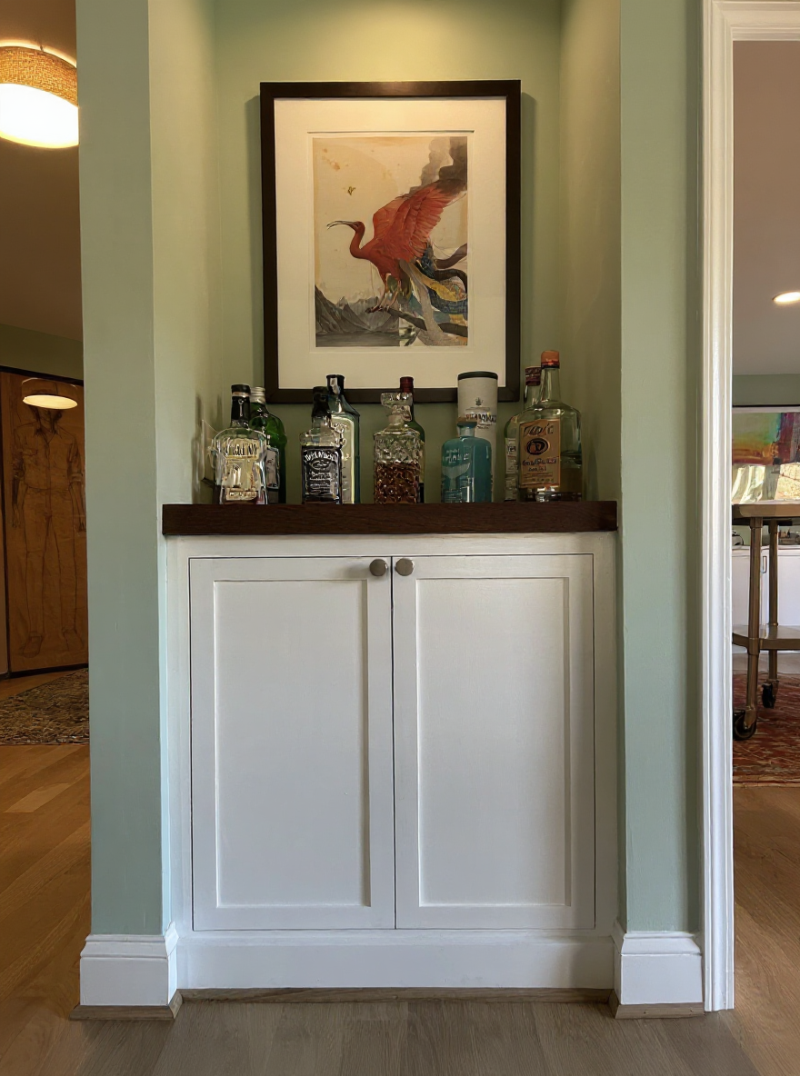A built-in cabinet with a dark brown countertop, displaying various bottles of alcohol, topped with a framed artwork of a bird with outstretched wings, set against a green wall inside a home.