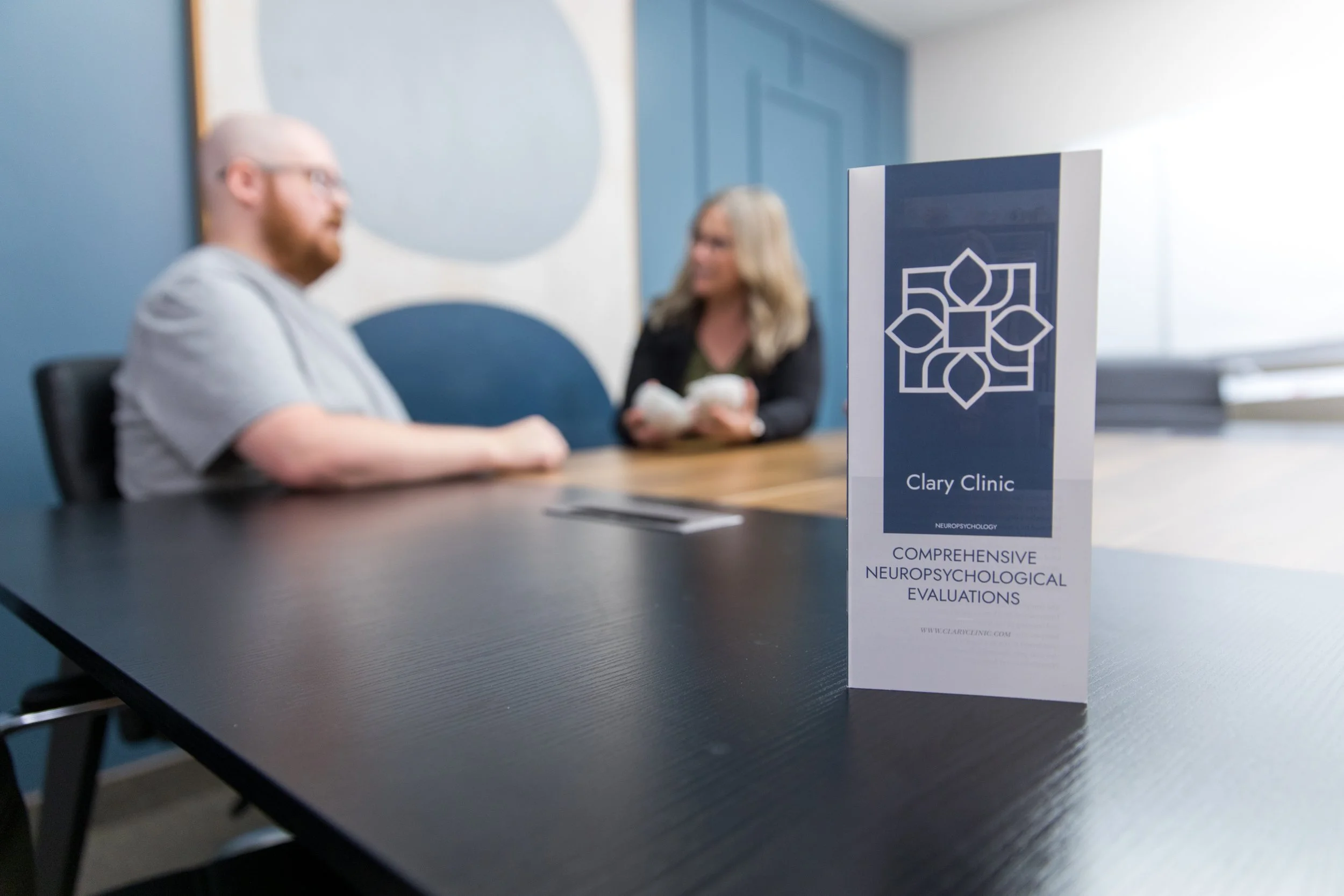 A pamphlet on a table reads 'Clary Clinic, Comprehensive Neuropsychological Evaluations.' In the background, a man and a woman are sitting at a conference table, blurred, engaged in conversation.